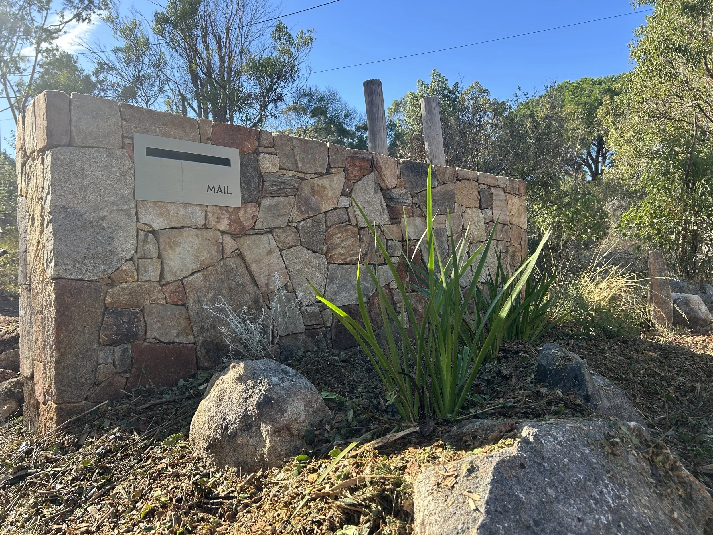 Stone wall with mail slot, surrounded by rocks and vegetation under a clear blue sky.