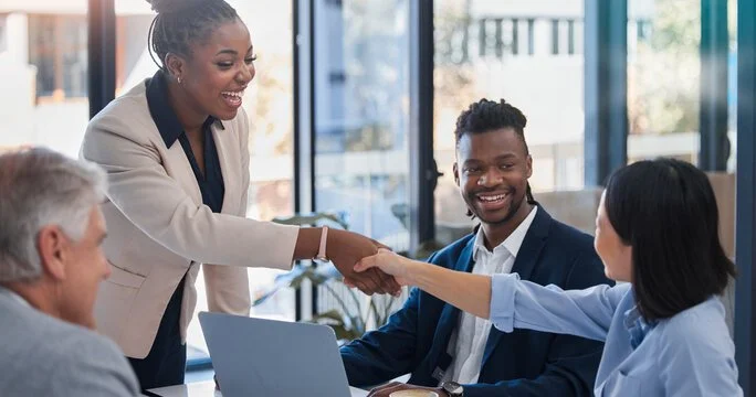 Businesswoman shaking hands with a woman in a business suit during a meeting, with three people sitting at a table in a modern office.