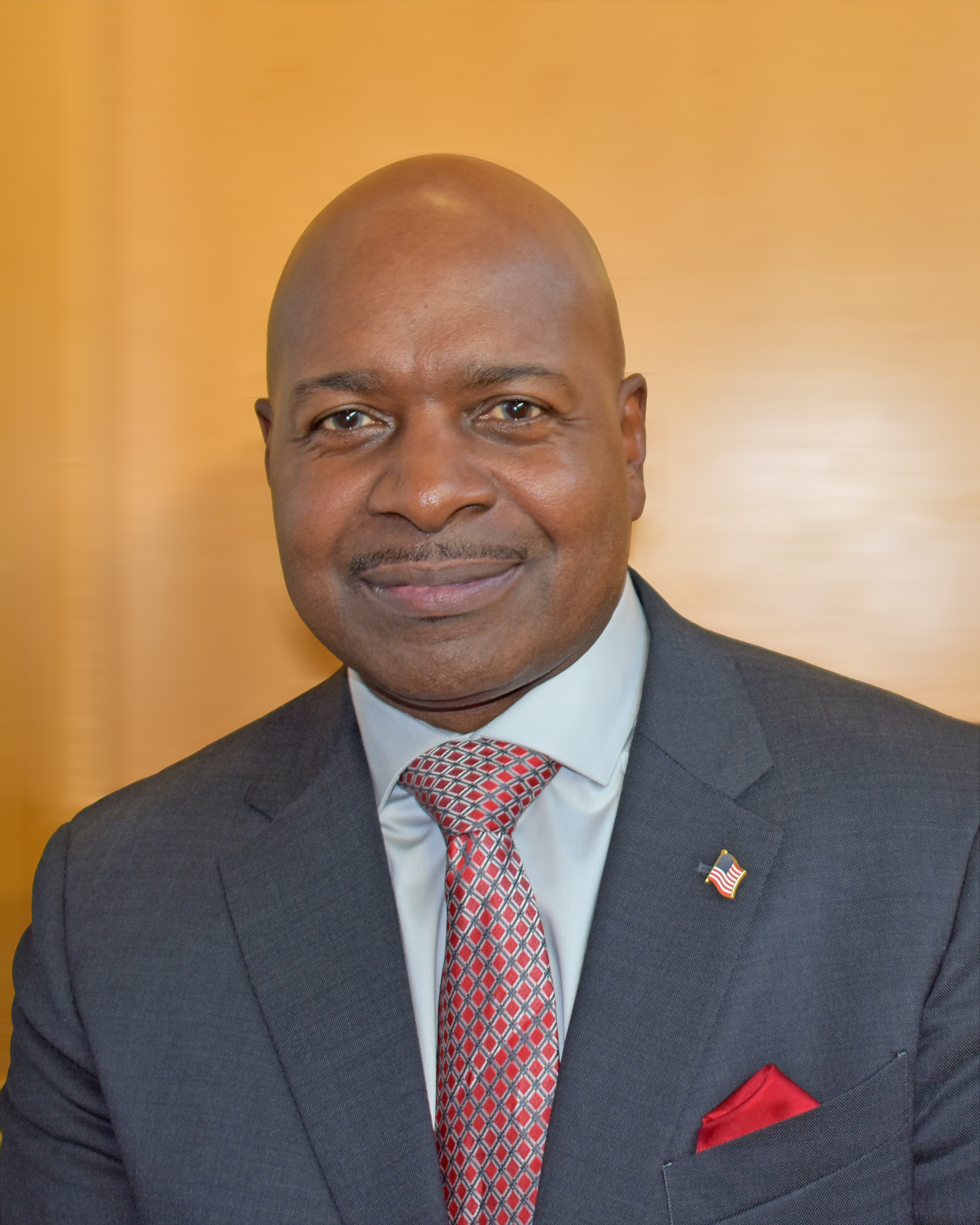 A man dressed in a suit and tie, with an American flag pin on his lapel, standing in front of a wooden background.