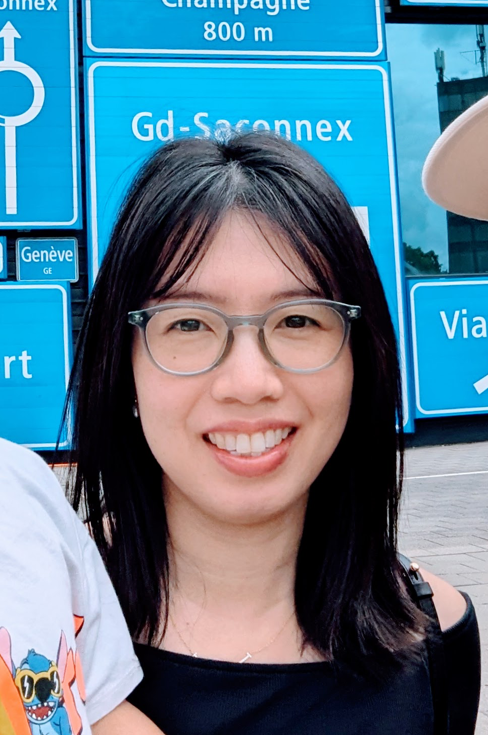 A woman with black hair, glasses, and a black top, smiling in front of blue road signs in Geneva, Switzerland.