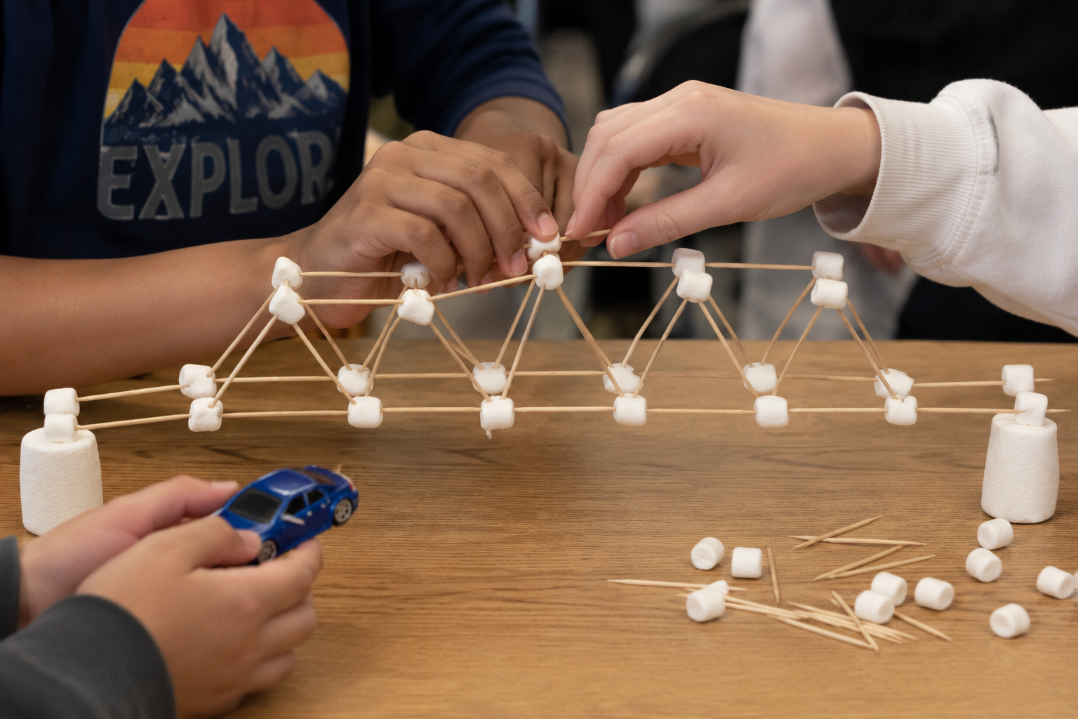Children building a bridge with marshmallows and toothpicks on a wooden table, with a toy car placed underneath.