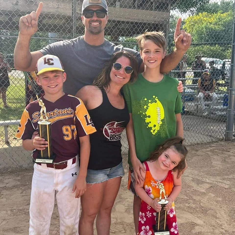 Group of five people, two adults and three children, at a baseball field. One boy is holding a trophy, and a girl is holding a smaller trophy. They are smiling and posing for the camera.