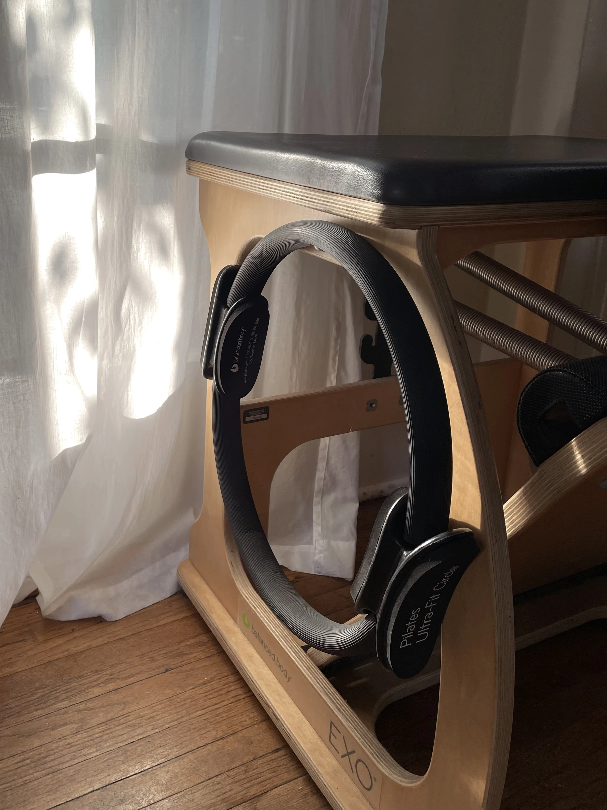 Wooden Pilates reformer with black padded footrest and springs, positioned near a white curtain and wooden floor.