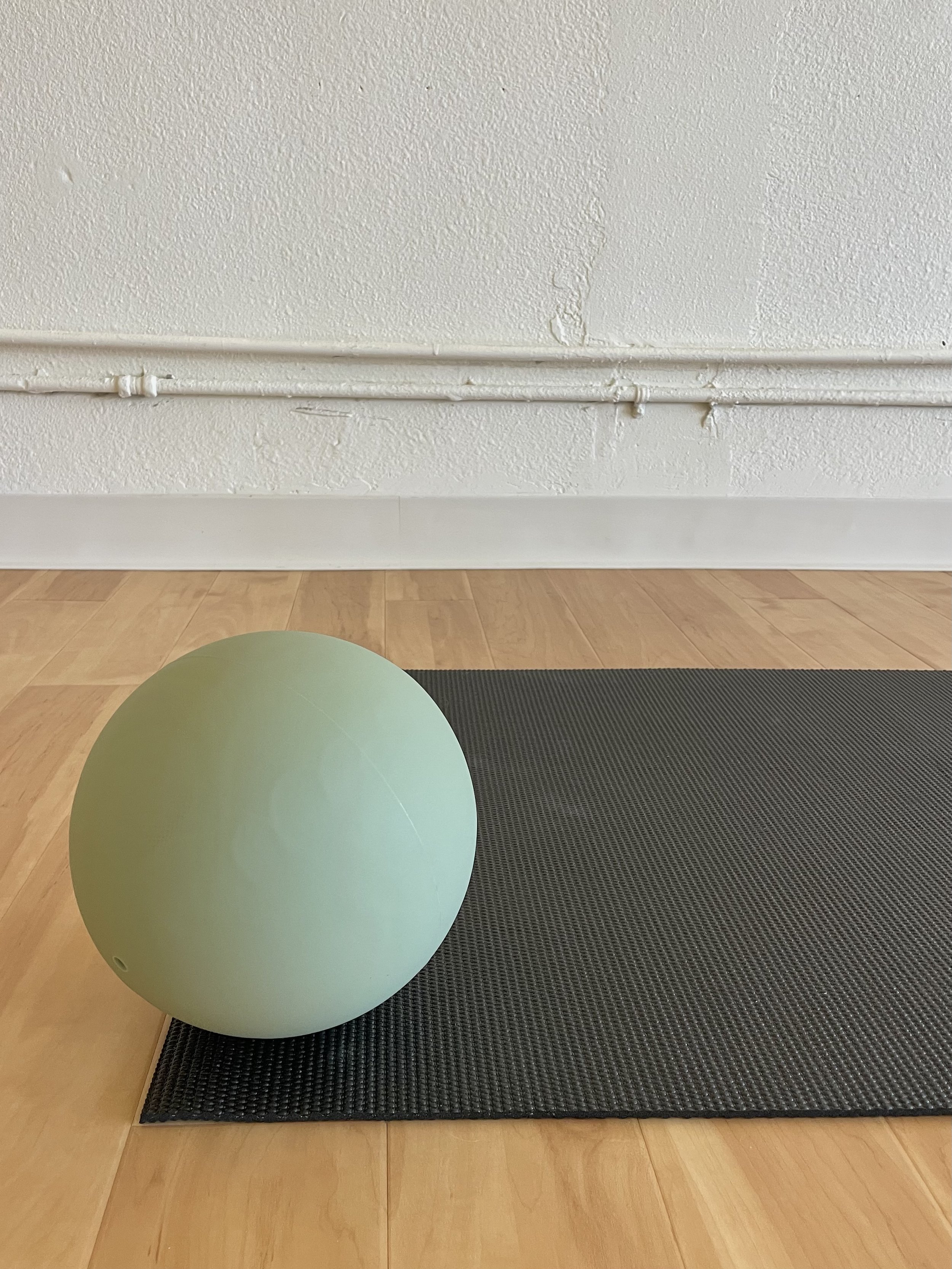 A green exercise ball resting on a black yoga mat on a wooden floor in front of a textured white wall with a white baseboard and pipes.