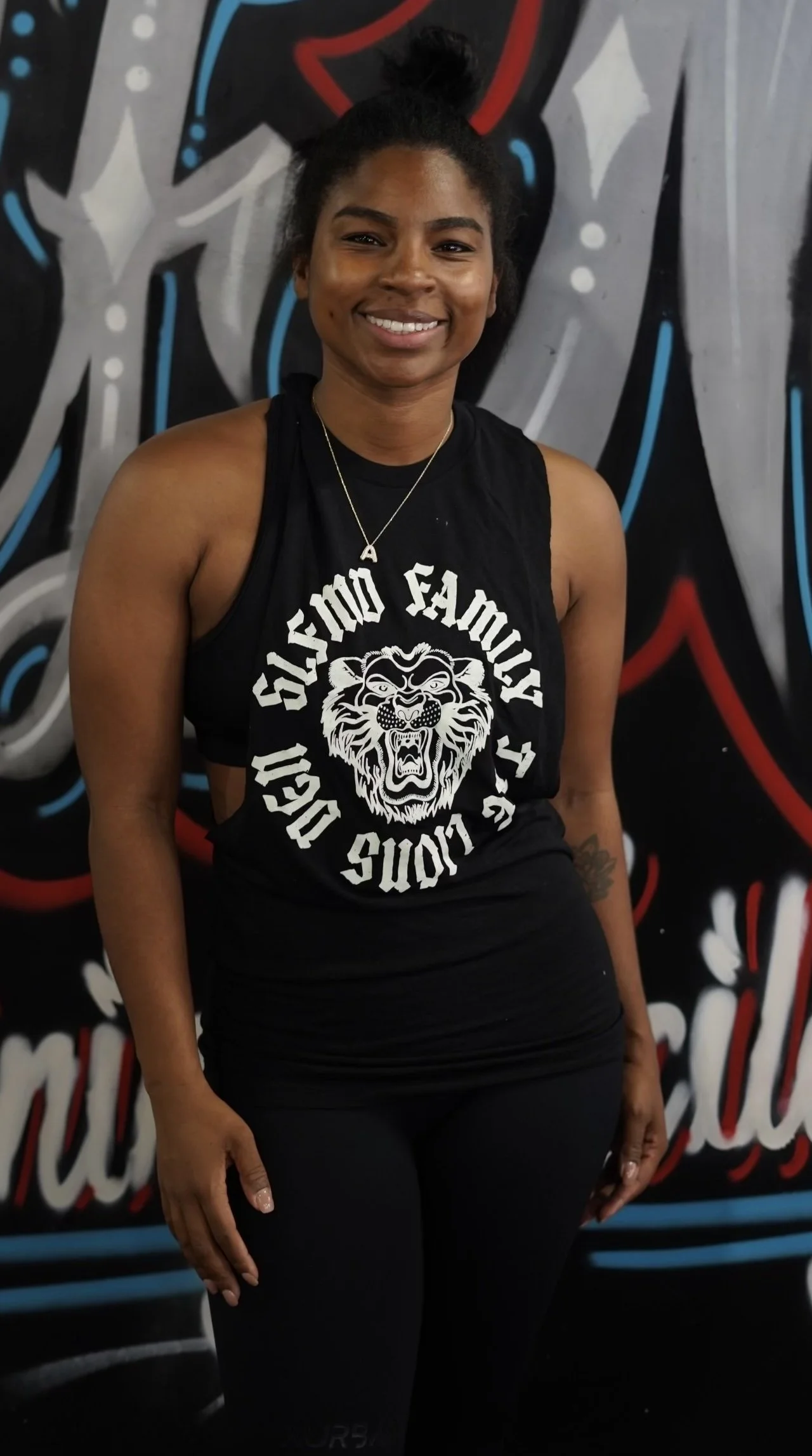 A woman smiling, wearing a sleeveless black shirt with a lion face and text, and standing in front of a graffiti wall with abstract designs and lettering.