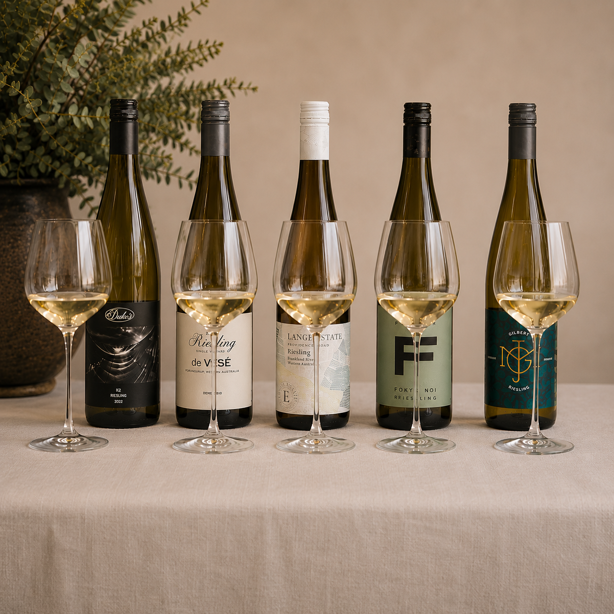 Five bottles of wine with glasses filled with wine in front of each bottle on a beige table.