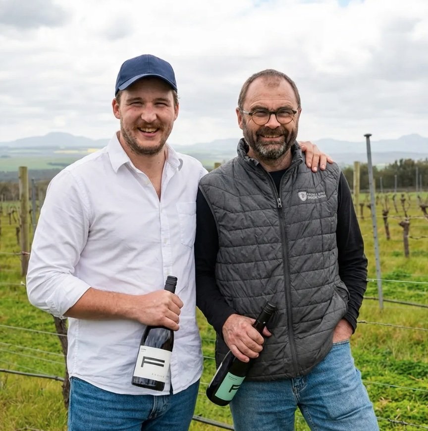 Callum and Mike standing together in a vineyard, each holding a bottle of Fervor wine.