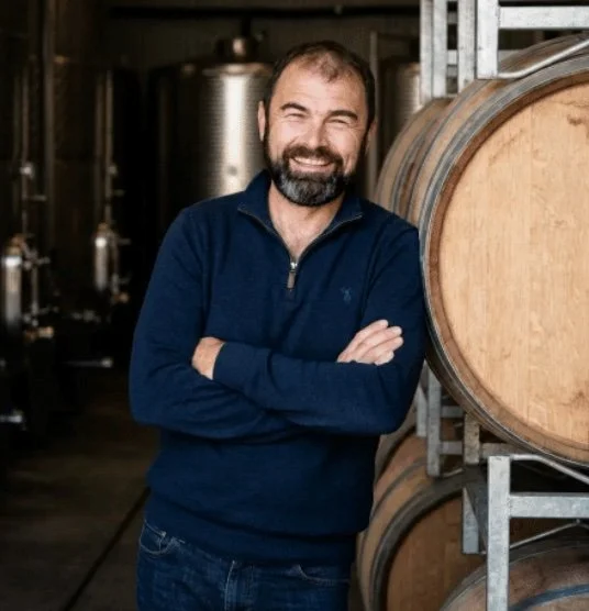 Mike standing with folded arms beside oak barrels in the Fervor Wines winery.