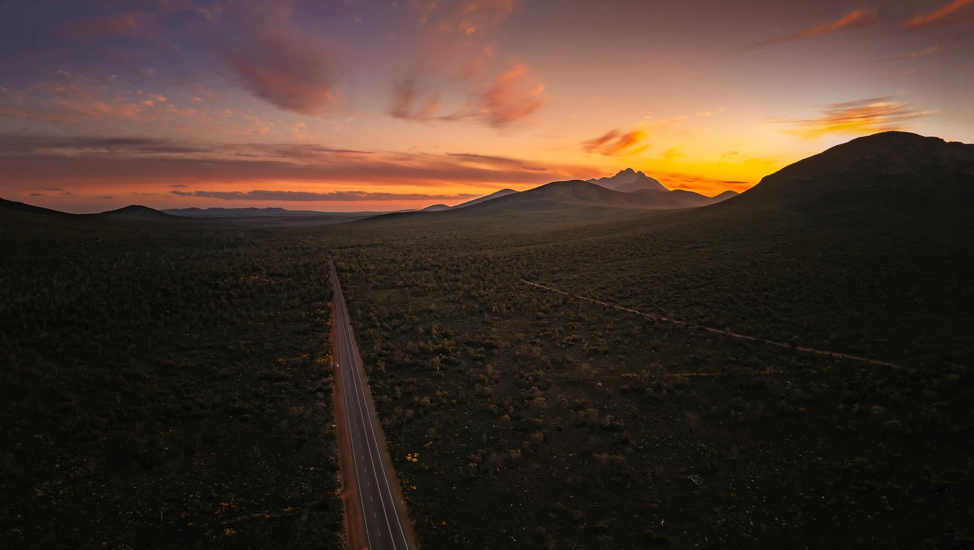 A scenic sunset over mountains with a road running through a dark, open landscape.