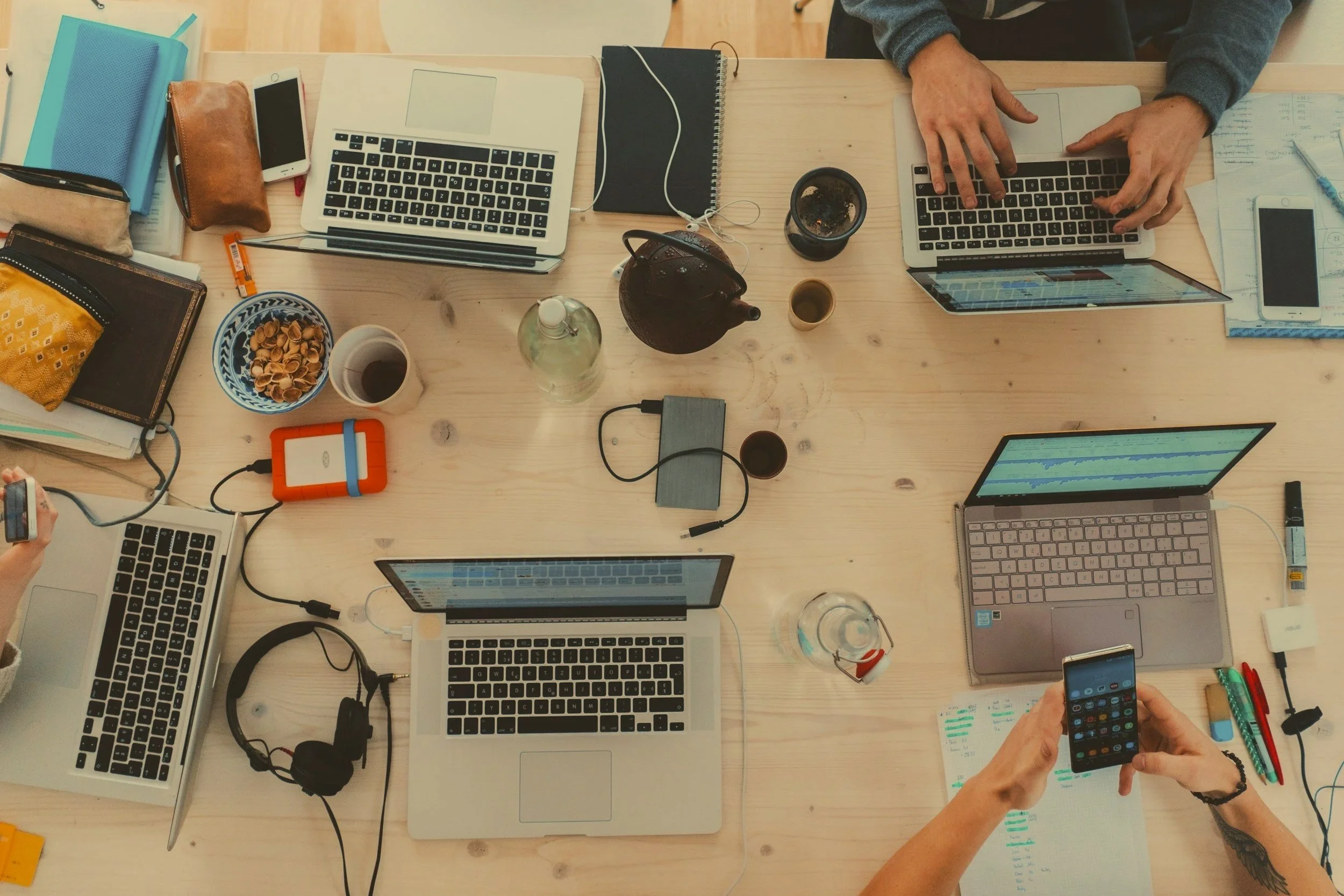 Top-down view of a workspace with multiple laptops, notebooks, phones, drinks, and personal items on a wooden table, with people using the devices.