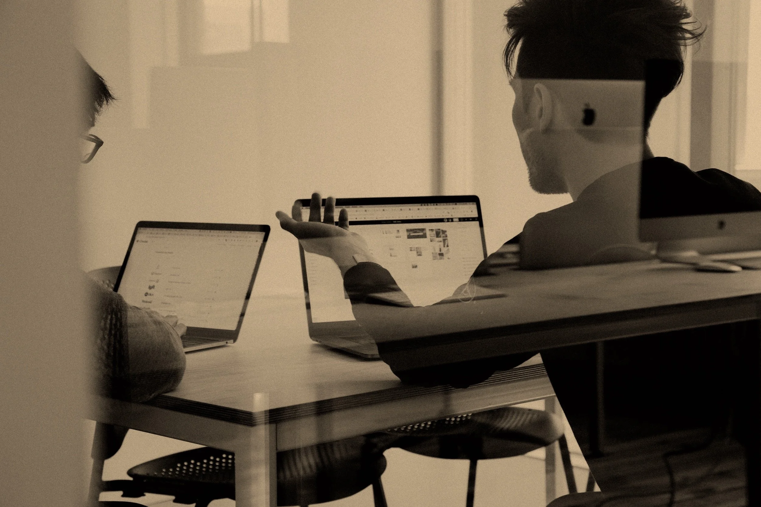 Two people sitting at a table with laptops in an office or meeting room, one person is gesturing towards the other's laptop screen.