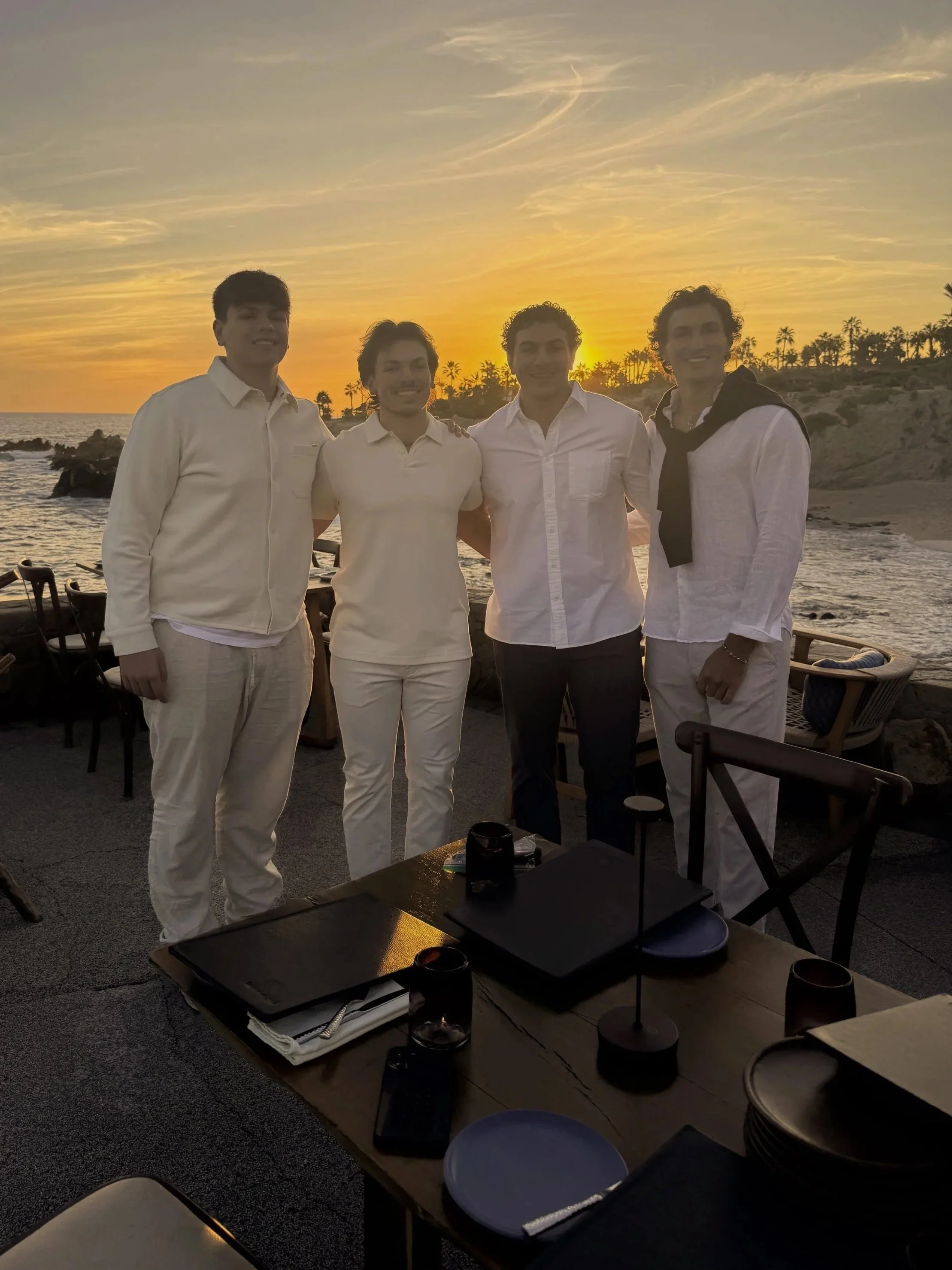 Four young men dressed in white and light-colored clothing stand together on a beach restaurant patio at sunset, with the ocean, rocks, and palm trees in the background.