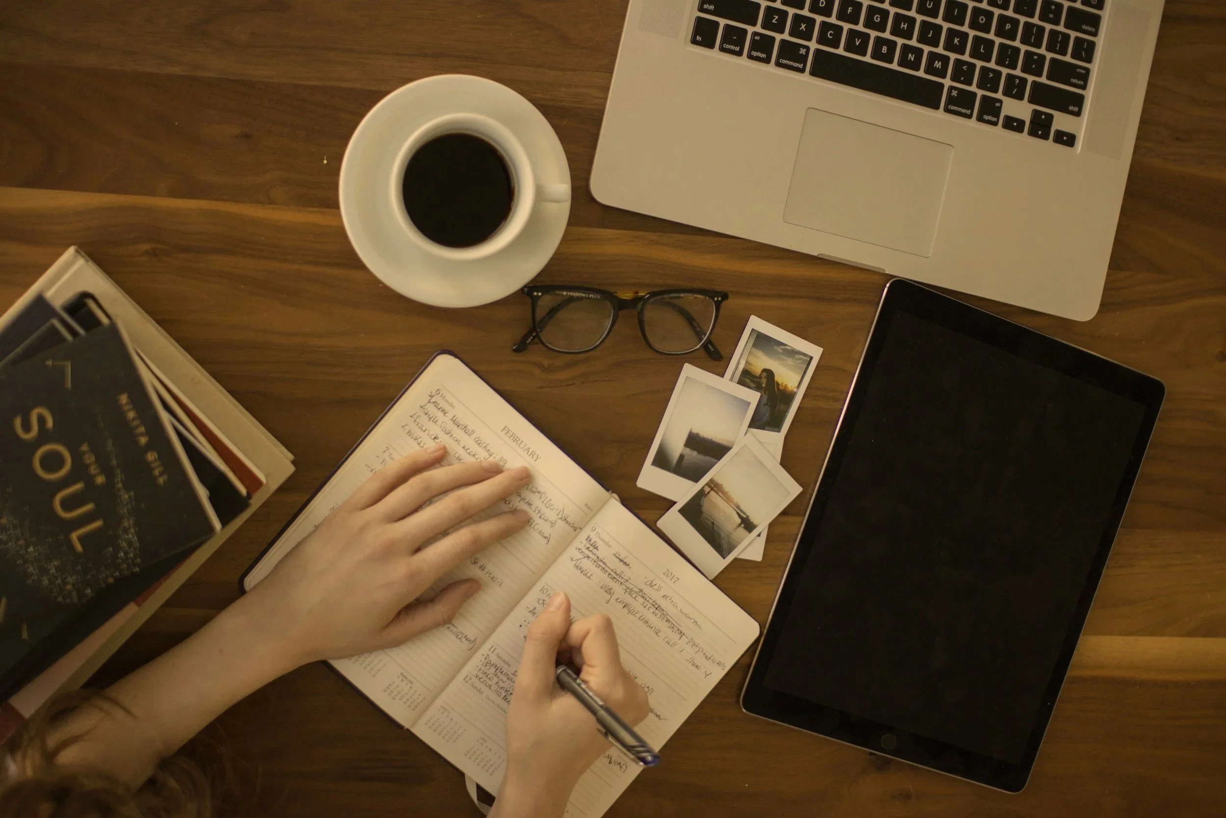 Overhead view of a workspace with a laptop, tablet, glasses, a cup of coffee, a planner, a person writing notes, a stack of books, and several printed photographs on a wooden table.