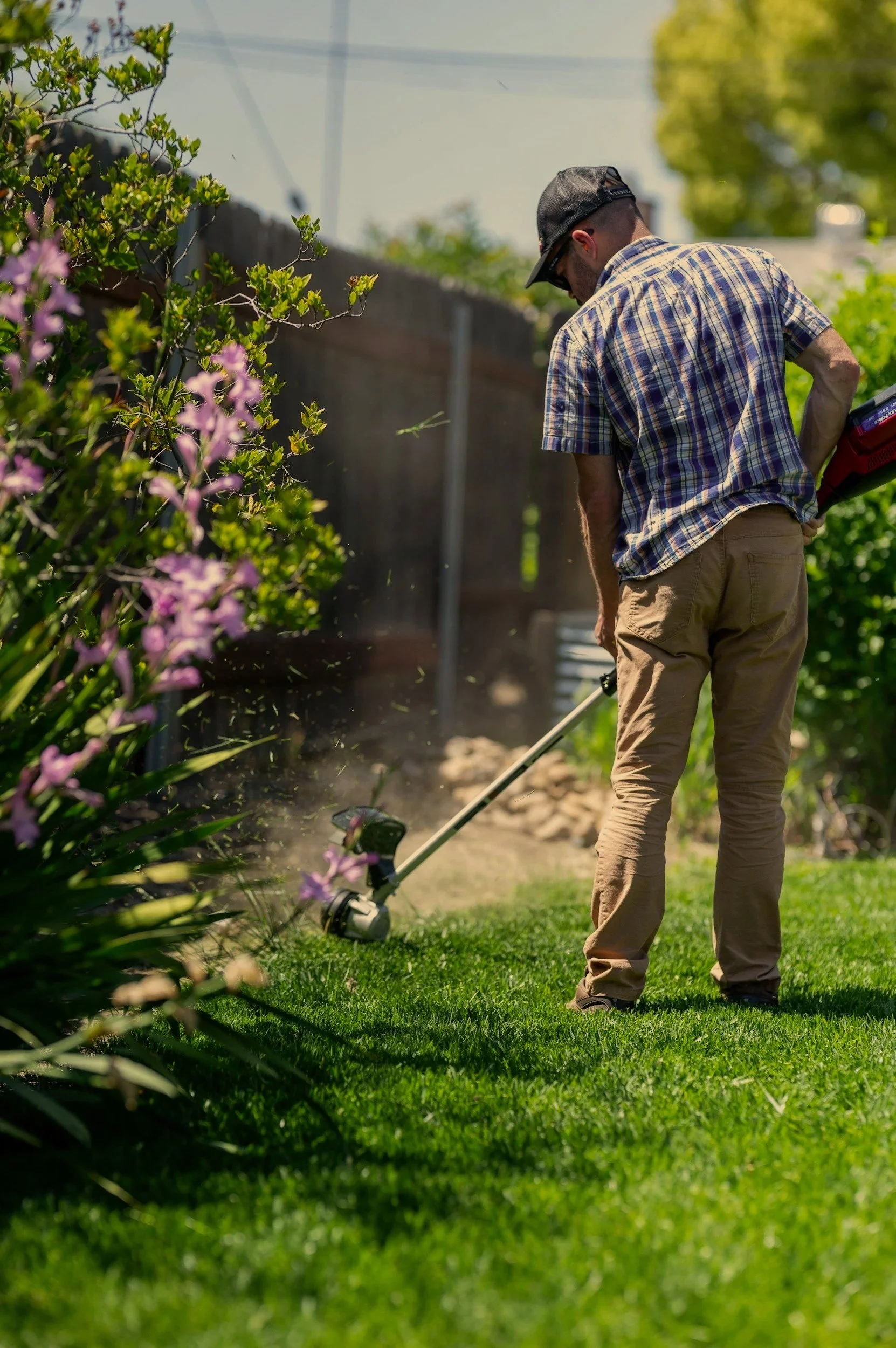 A man wearing a plaid shirt, tan pants, and a black cap trimming bushes with a grass trimmer in a backyard.