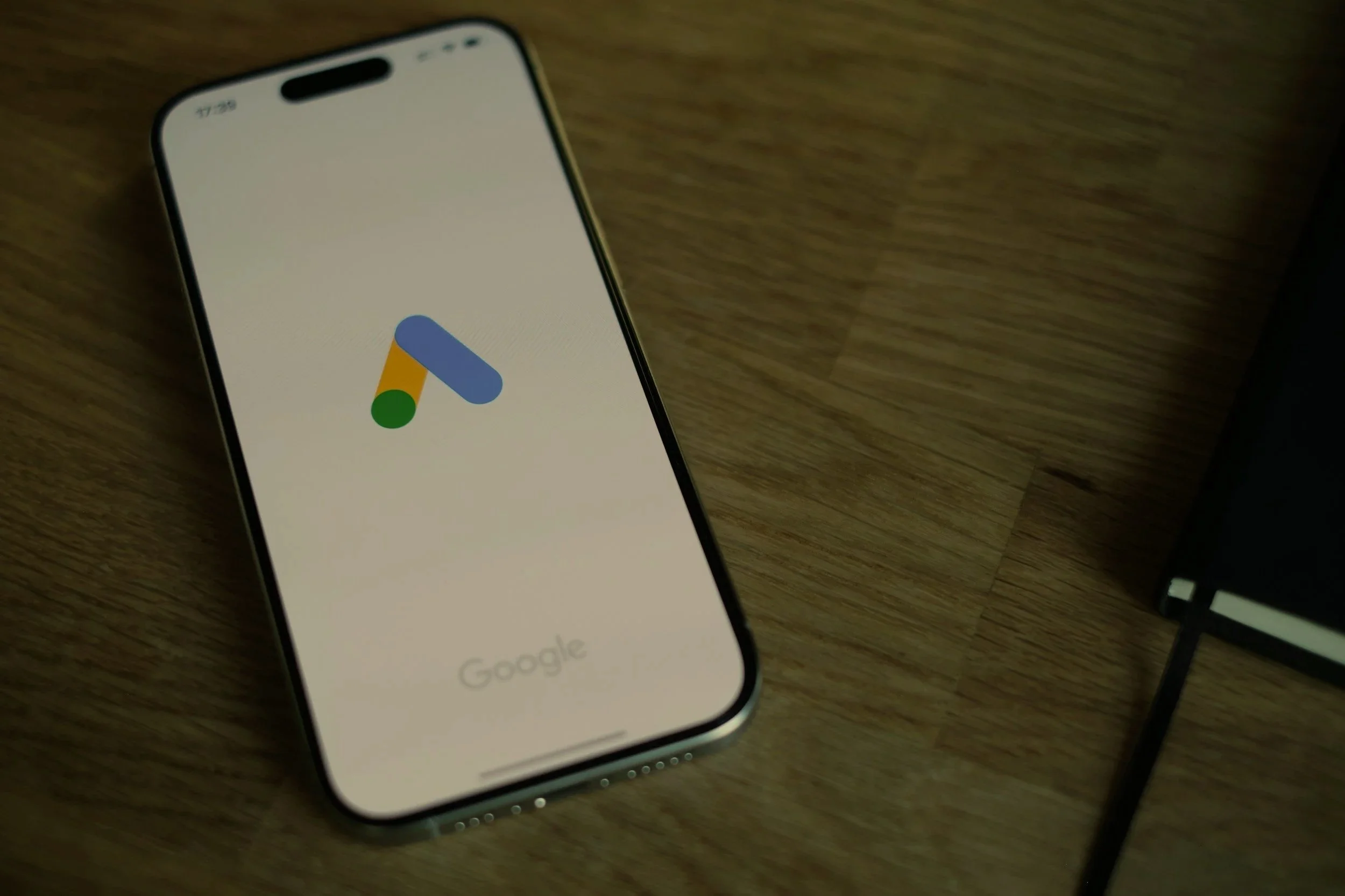 An iPhone with a Google Ads app logo on the screen, lying on a wooden surface next to a black notebook.