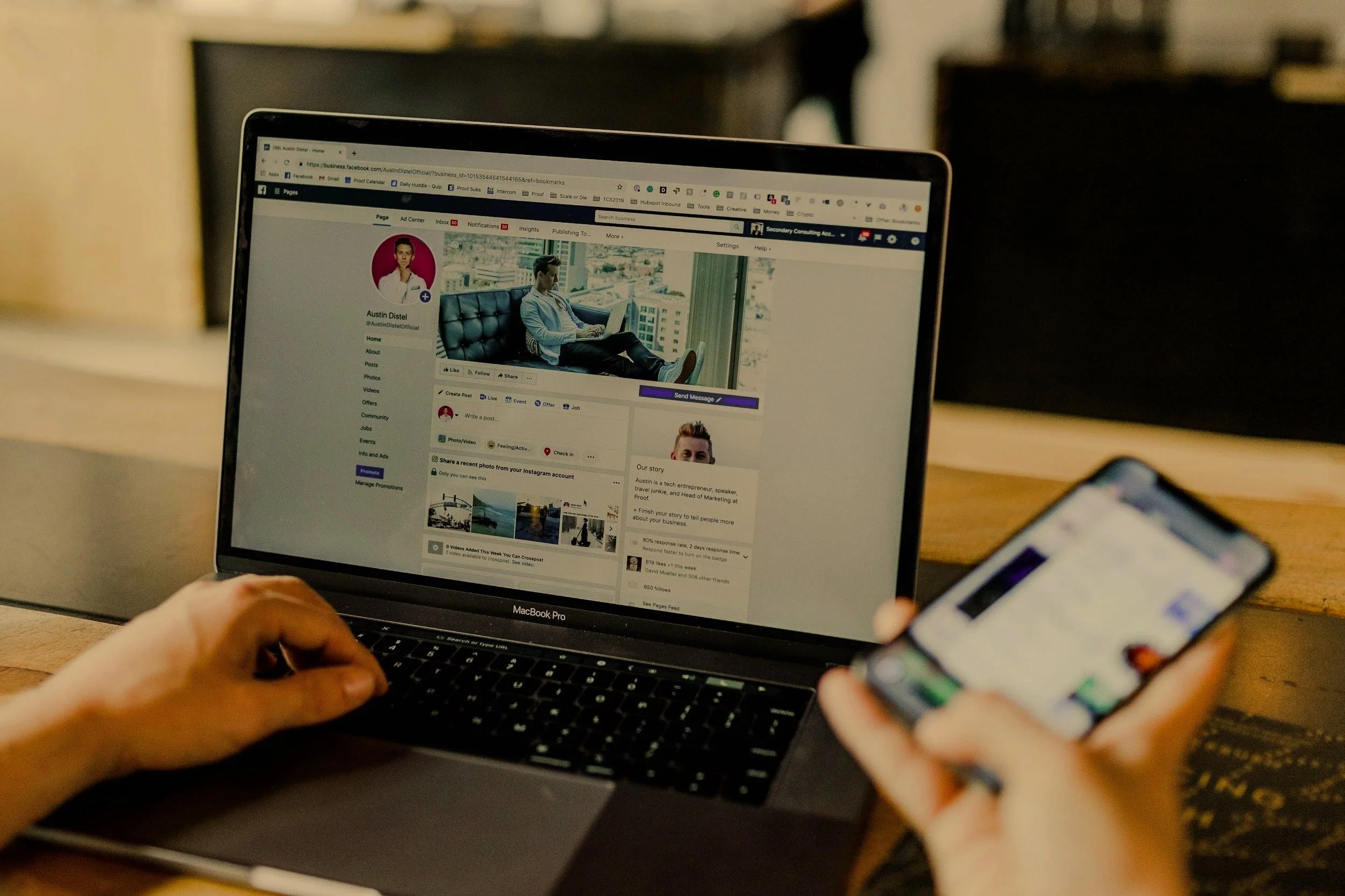 Person using a MacBook Pro and smartphone, browsing Facebook on the laptop with a wooden table and blurred background.