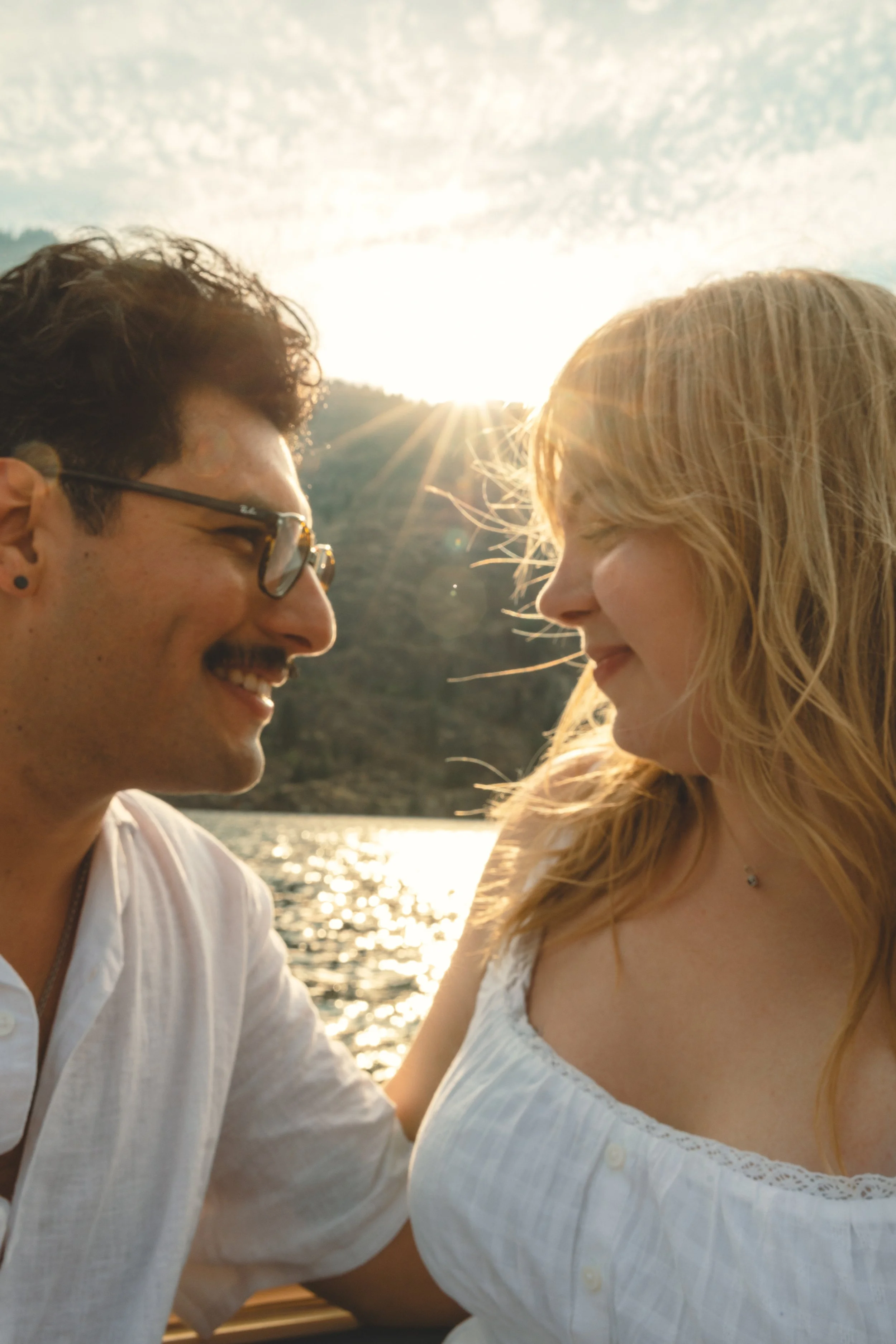 A man and a woman are close to each other, smiling and looking into each other's eyes, with the sun setting behind a mountain in the background.