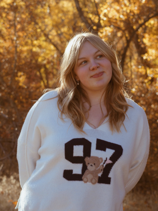 Young woman with blonde hair wearing a white sweater with the number 97 and a teddy bear on it, standing outdoors in a park during autumn.