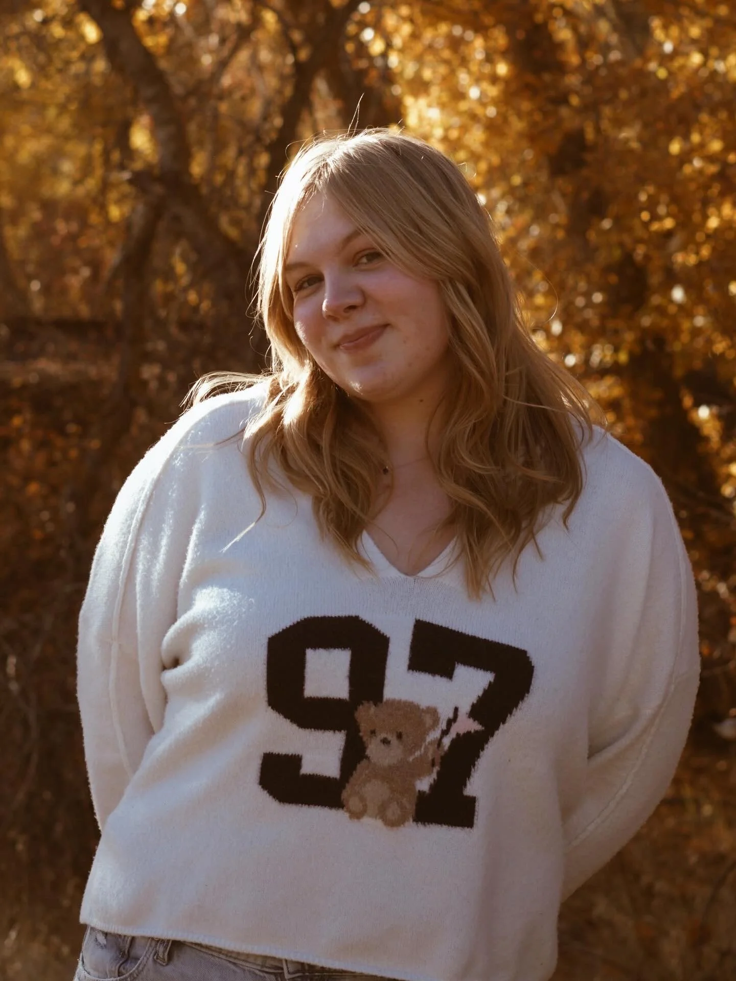 A woman with blonde hair wearing a white sweatshirt with a large black number 92 and a small illustration of a teddy bear on the front, standing outdoors with orange autumn leaves in the background.