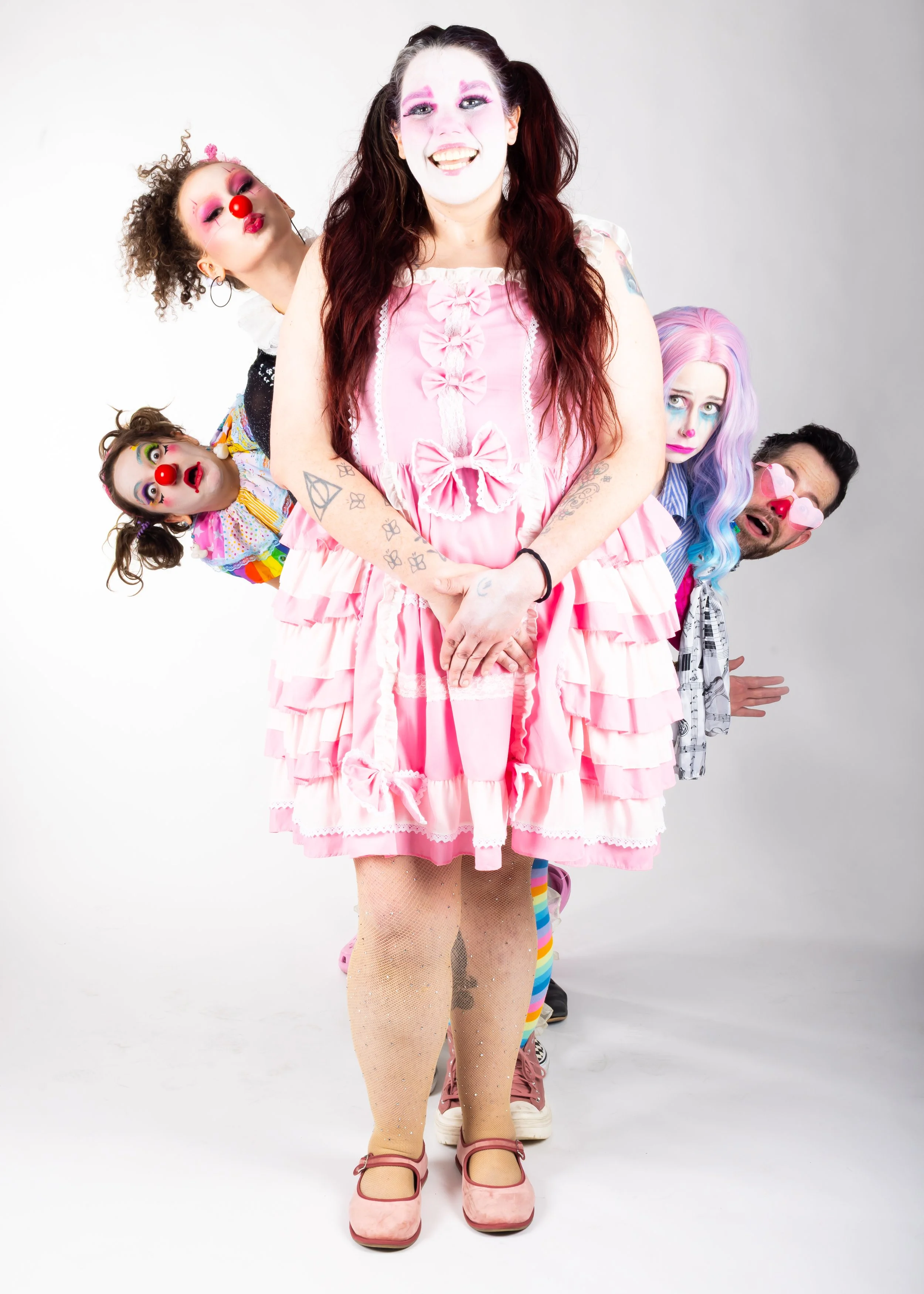 A group of four people dressed in colorful, eccentric costumes with clown makeup, posing together in front of a plain white background. The person in the front wears a pink dress, fishnet stockings, and pink shoes, with rainbow-colored socks. The second person is dressed as a clown with rainbow hair and makeup. The third person wears brightly colored makeup and a clown costume. The fourth person, partially visible, has dark hair, clown makeup, and a clown nose.