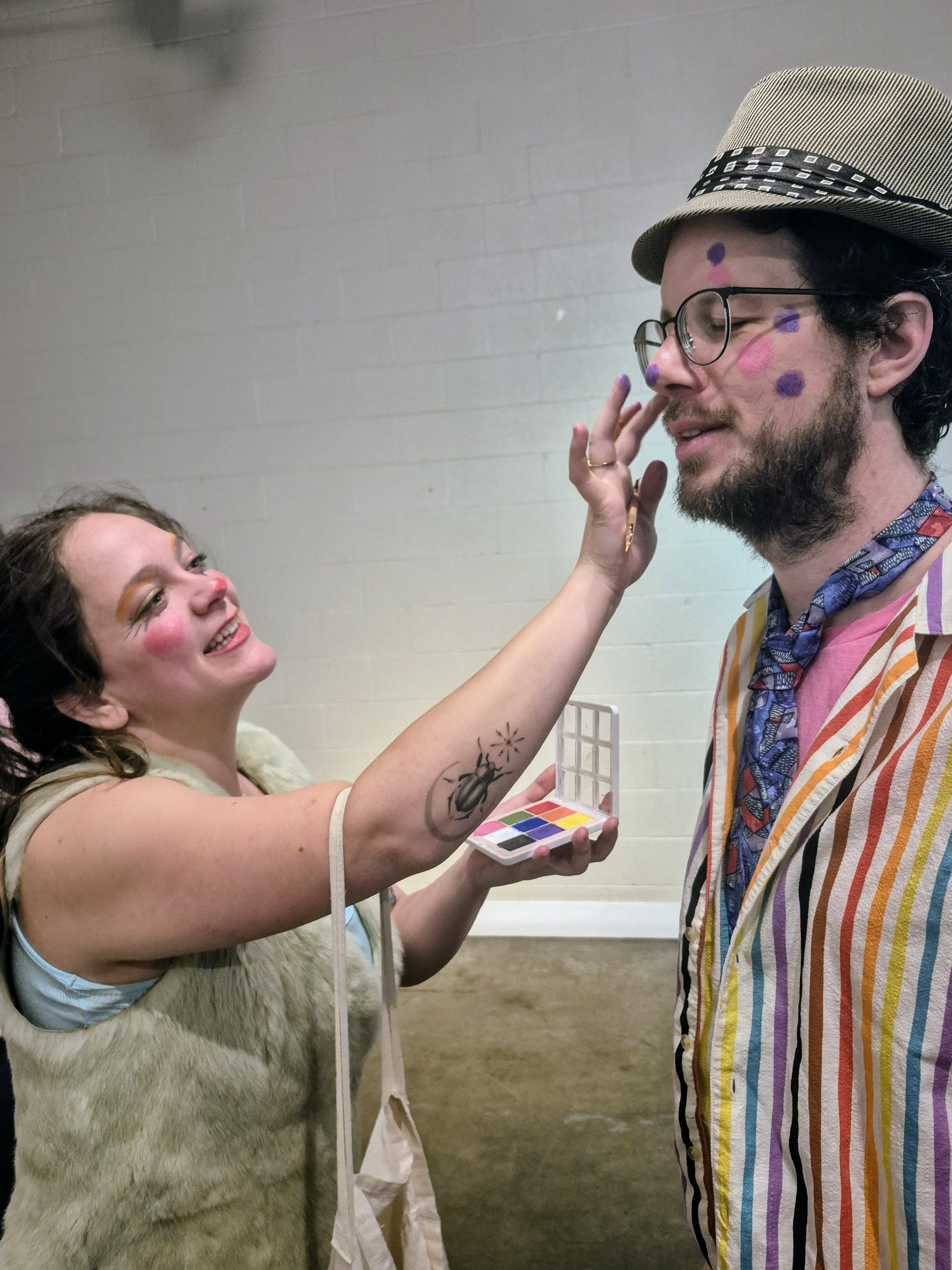 A woman with clown makeup applying face paint to a man's face, who is wearing striped clothing, a hat, and glasses, in a playful setting.