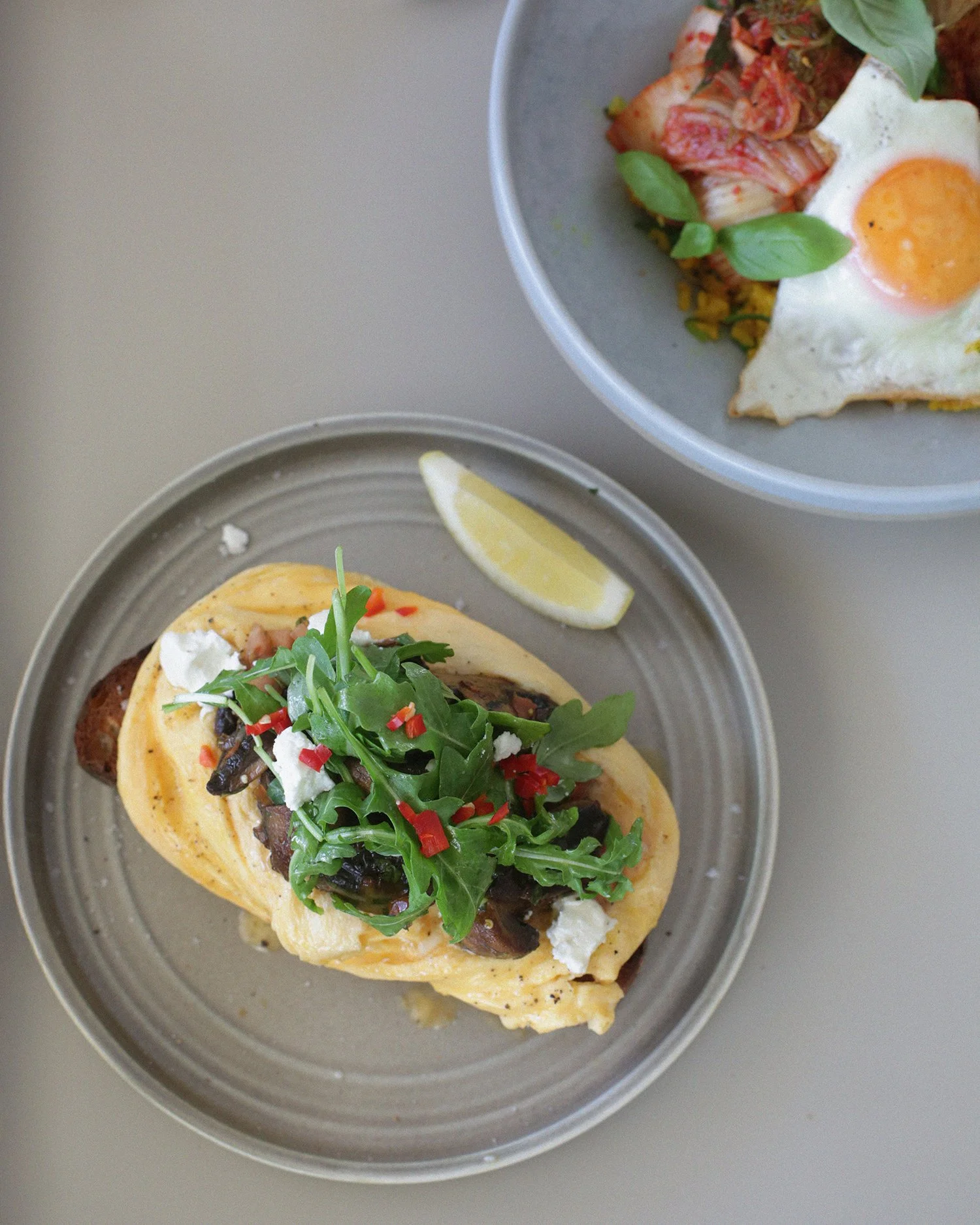 Eggs with a side of vegetables on a gray plate with a lemon wedge, and another dish with an egg and vegetables in a gray bowl.