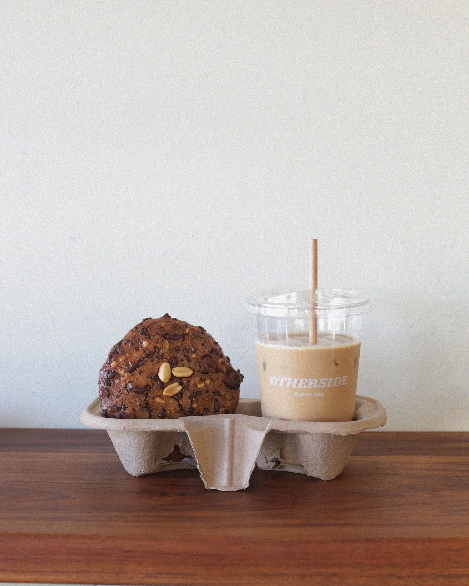 A tray with a chocolate chip cookie and a iced drink in a clear cup with a straw, on a wooden surface against a plain wall.