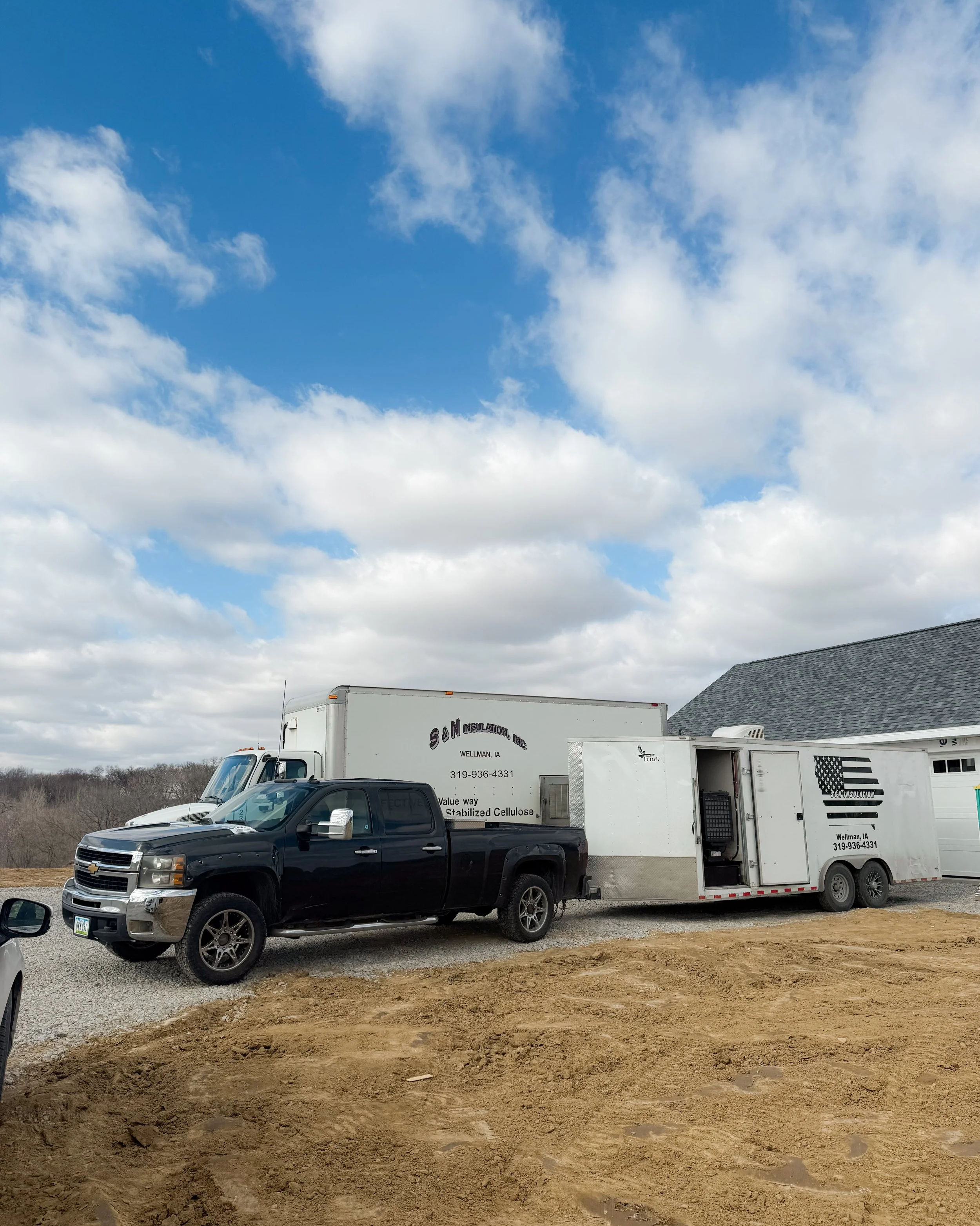A black pickup truck is parked in front of a white moving truck at a construction site with dirt and gravel in the foreground, and a blue sky with scattered clouds overhead.