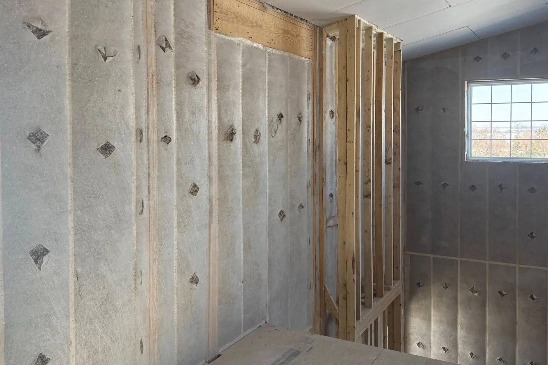 Interior view of a room under construction, showing wall studs and drywall with a window in the background.