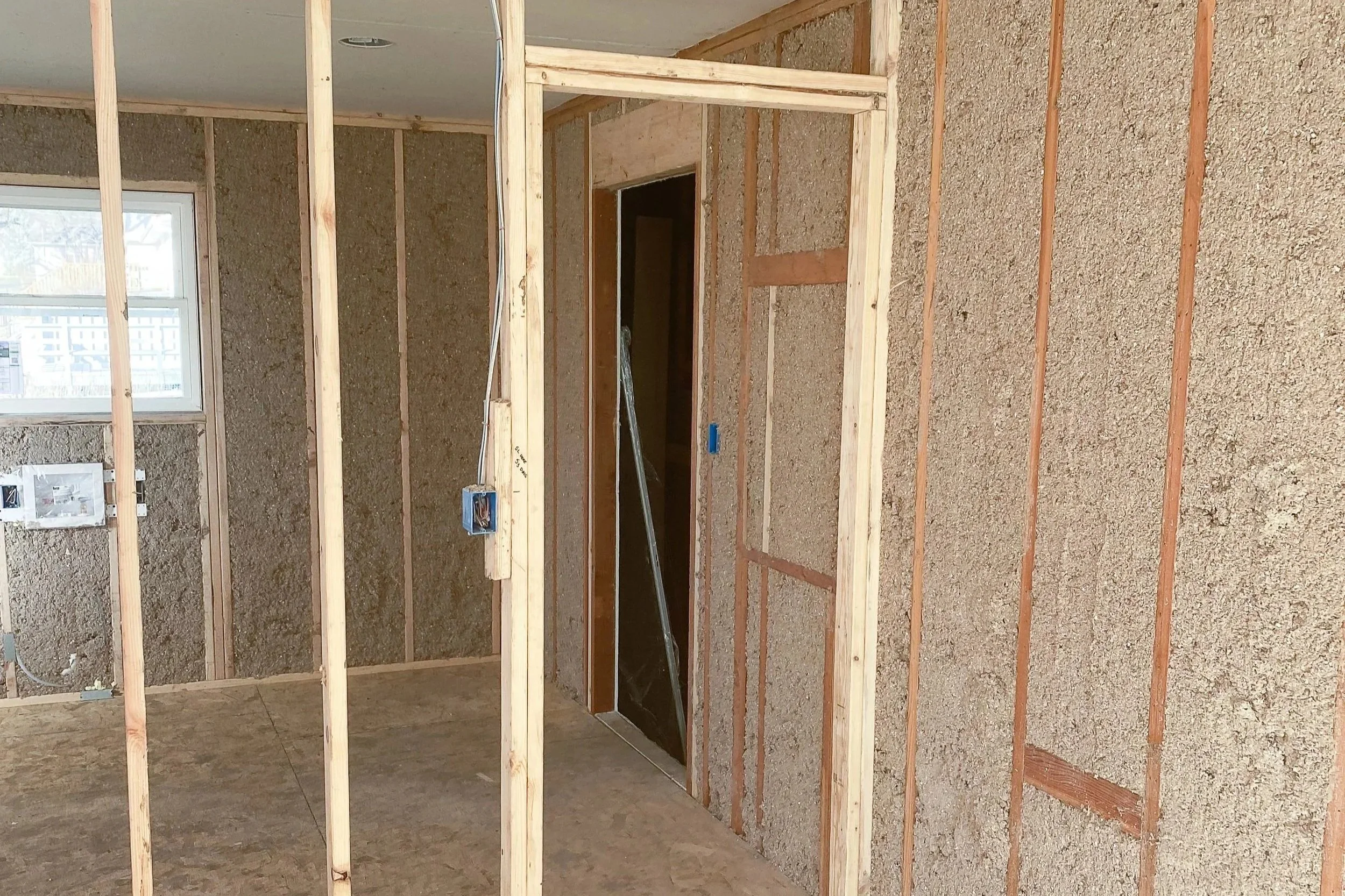 Interior of a house under construction with exposed wooden framing and drywall. There is an open doorway and a window with a view outside.