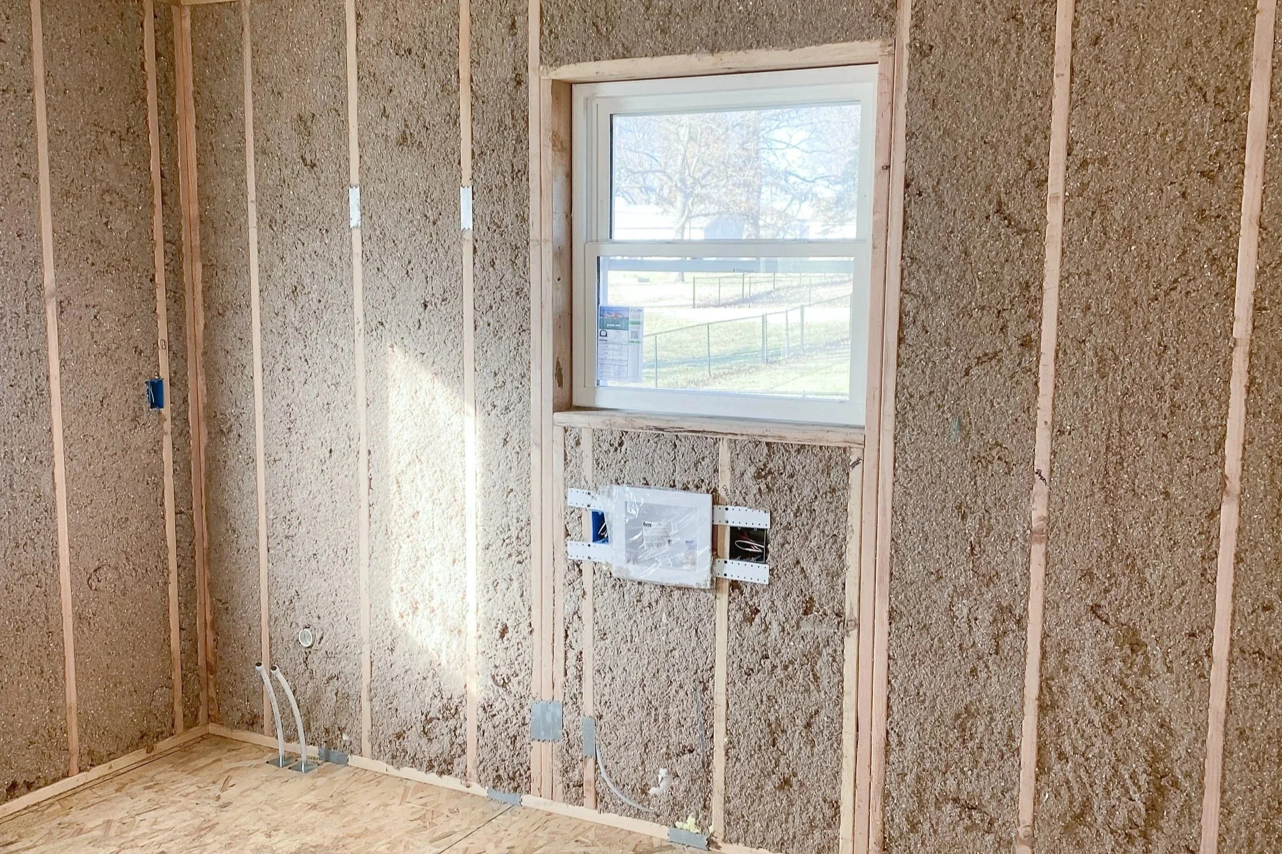 Interior of a room under construction with exposed wall studs, insulation, and a single window showing an outdoor scene with trees and a fence.