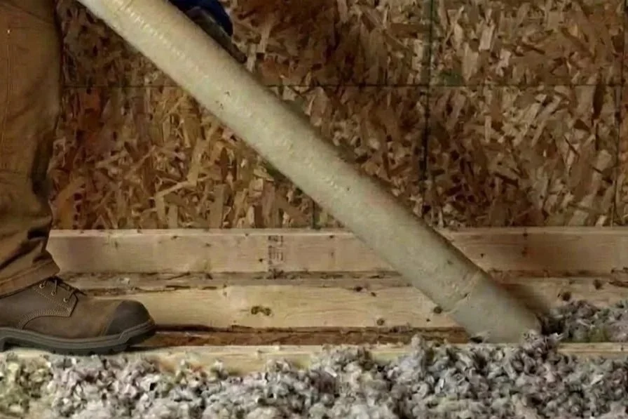 Close-up of a person's boot and leg, standing on a wooden floor with sawdust, next to a slanted PVC pipe resting on the floor inside a wooden and plywood shed or construction area.