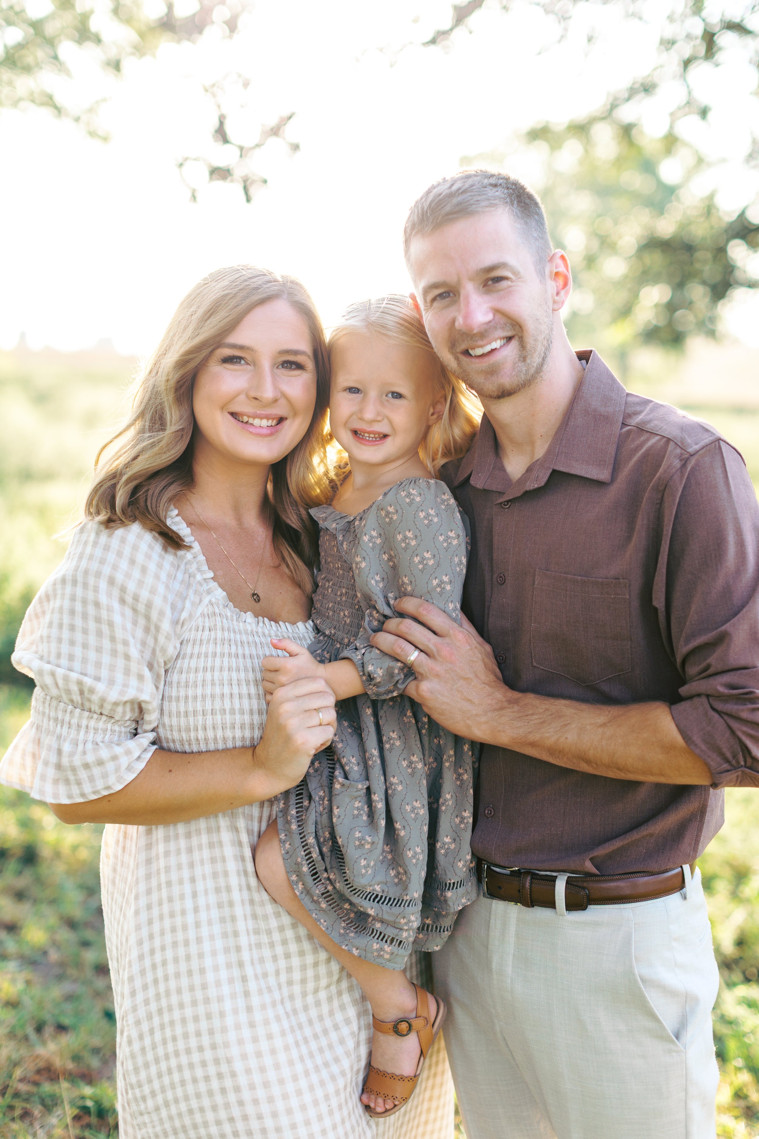A happy family of three outside in a park on a sunny day. The family includes a woman, a man, and a young girl, all smiling at the camera.