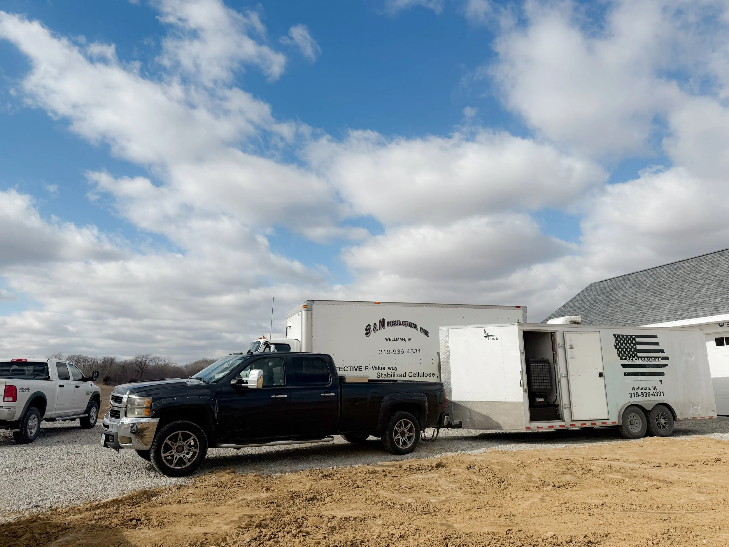 Vehicles and a trailer parked on a gravel lot under a partly cloudy sky.