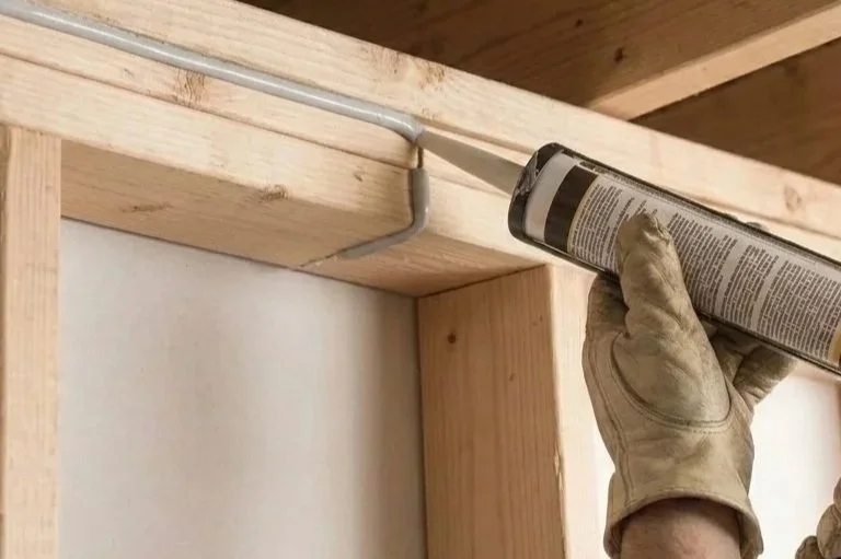 A person wearing a work glove applying caulk along the corner between a wooden frame and a wall.