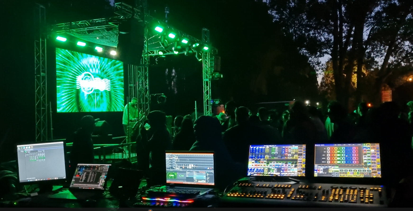 Conjunto de equipos de producción y sonido en un evento musical con una pantalla grande y un escenario iluminado con luces verdes y púrpuras.