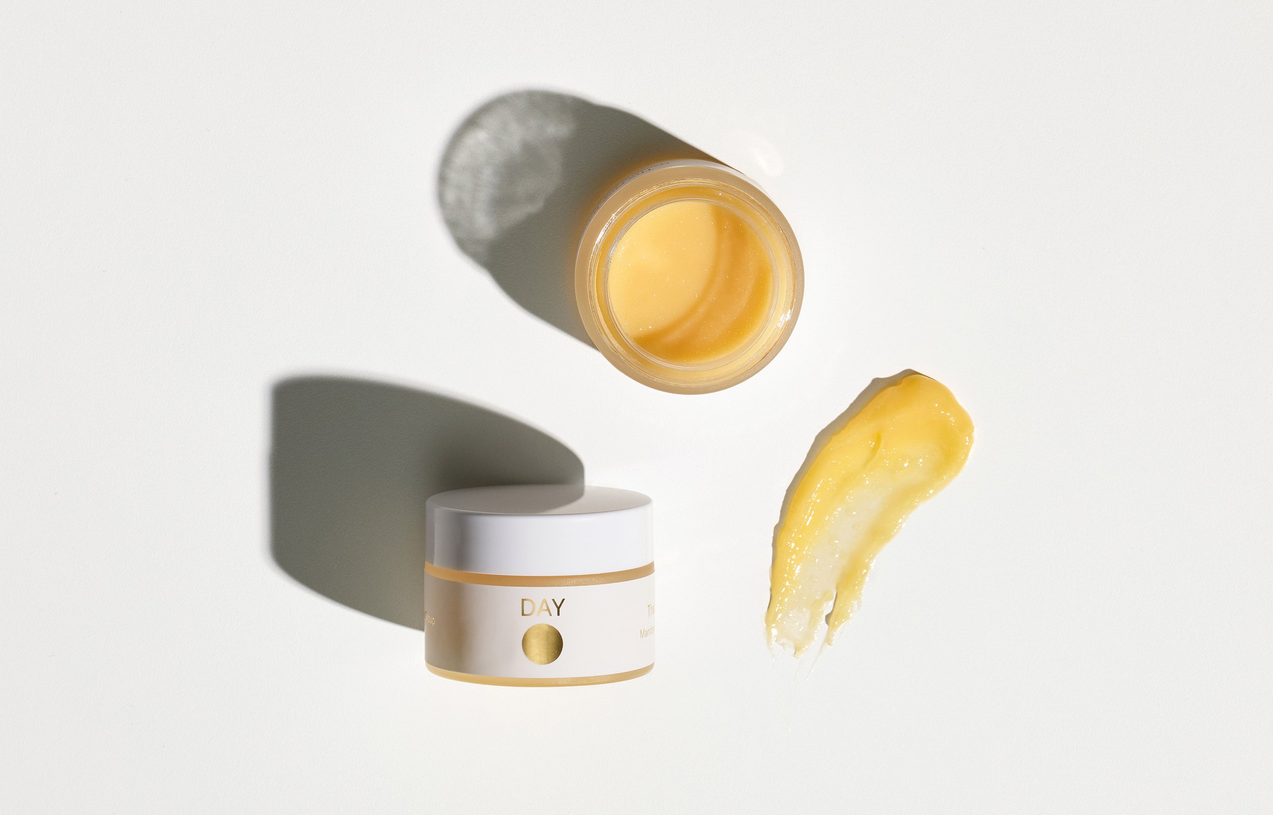 A white jar labeled 'DAY' with a gold circle, an open small container with yellow cream inside and a swatch of yellow cream on a white surface, casting shadows.