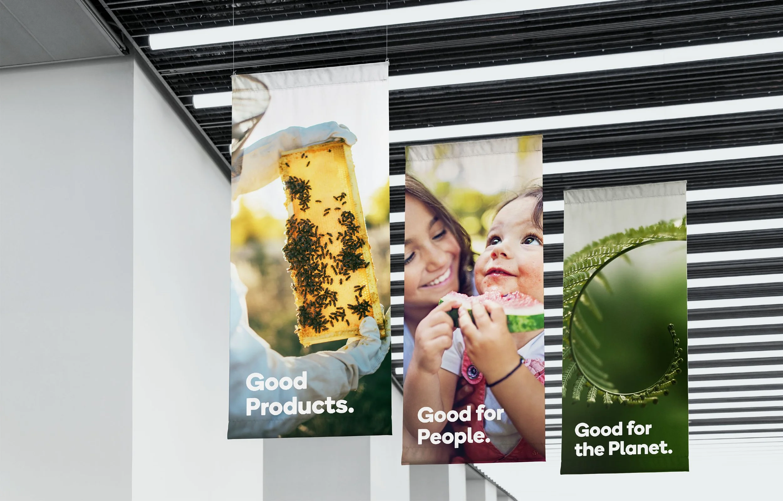 Three vertical banners hanging from the ceiling, each with a different environmental or social message, displaying images of bees on honeycomb, a woman and a child enjoying a snack, and a close-up of a fern leaf.