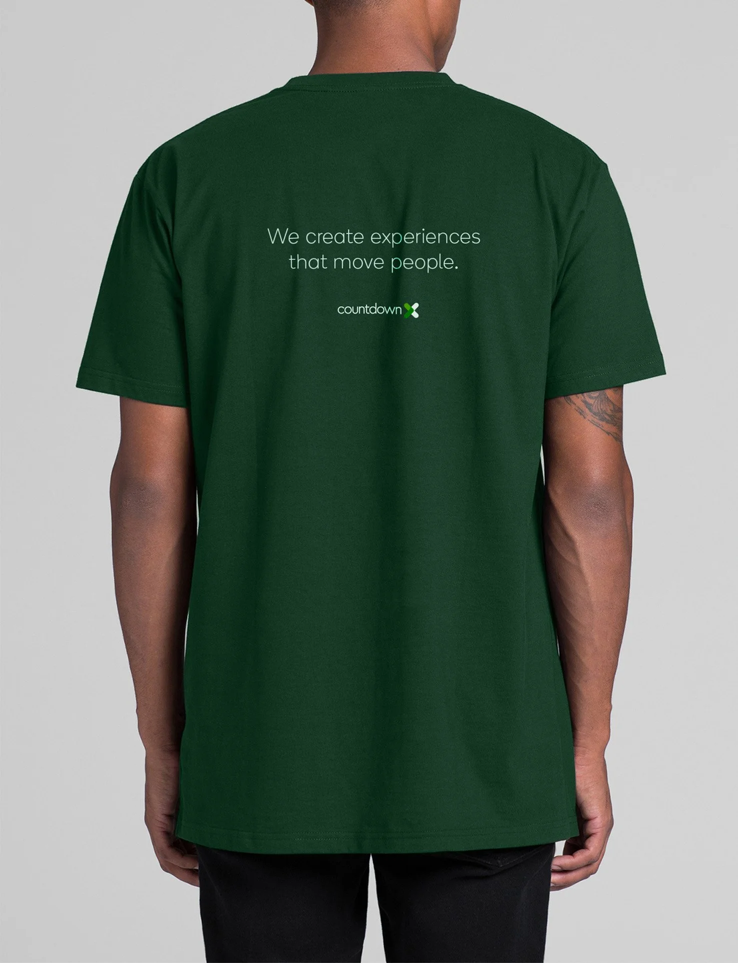 Back of a person wearing a dark green t-shirt with the text "We create experiences that move people" and a small logo reading "Countdown" on it.