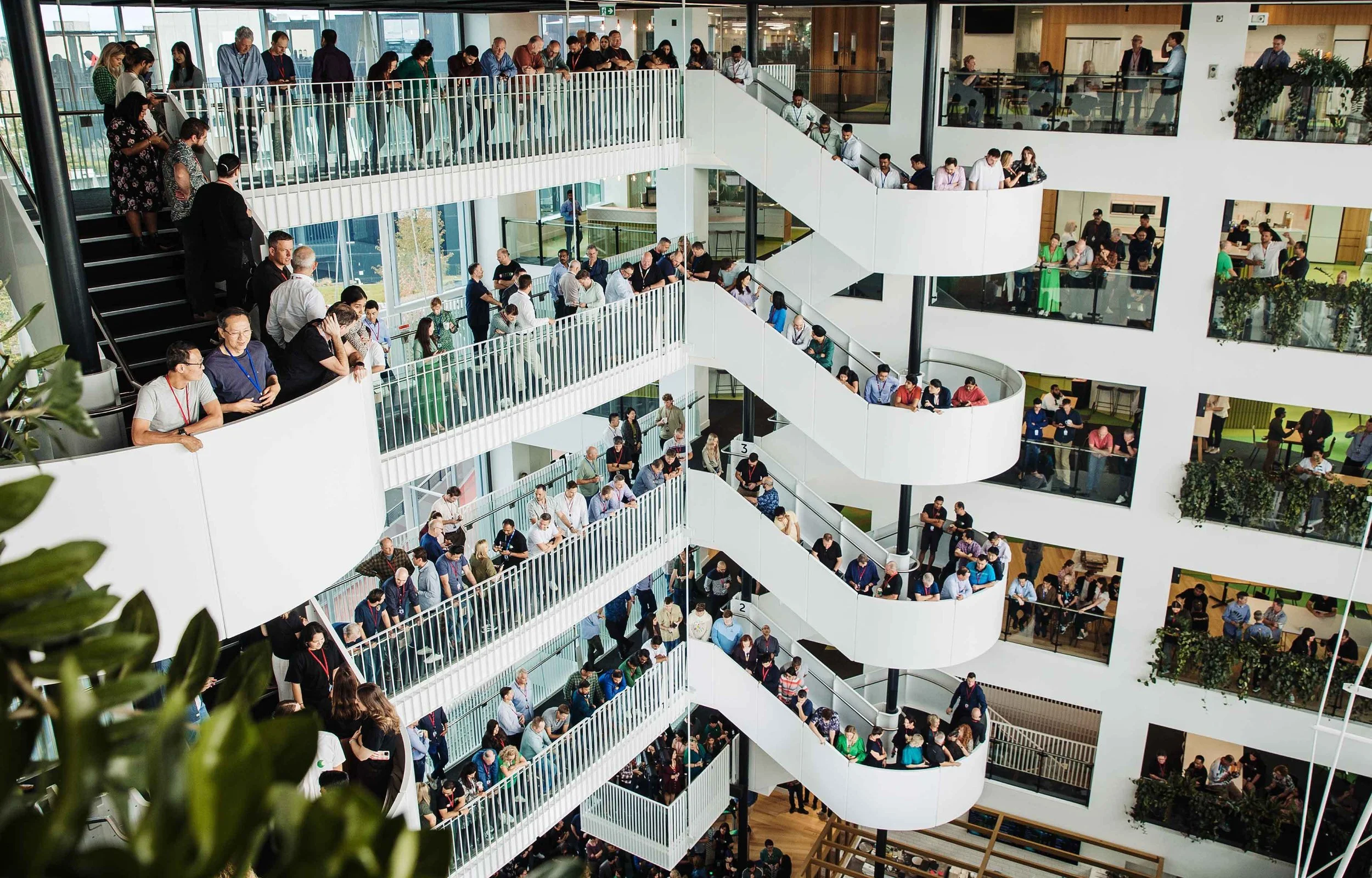 Multiple floors of a modern office building filled with people standing in long lines along white safety railings, waiting on staircases and on balcony walkways.
