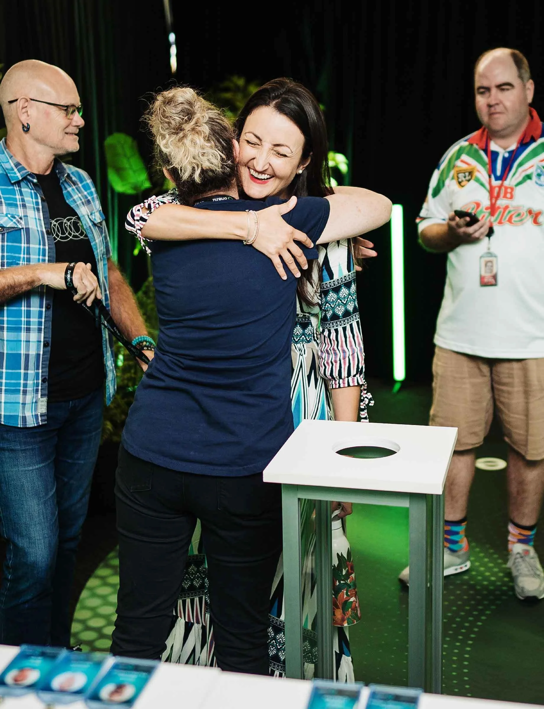 Two women hugging each other warmly in a celebratory setting. One woman has long dark hair and is smiling, while the other has curly blonde hair. There are three men in the background, one with glasses and a plaid shirt, and another in a colorful sports jersey. The environment suggests an event or party with a dark background and green lighting.