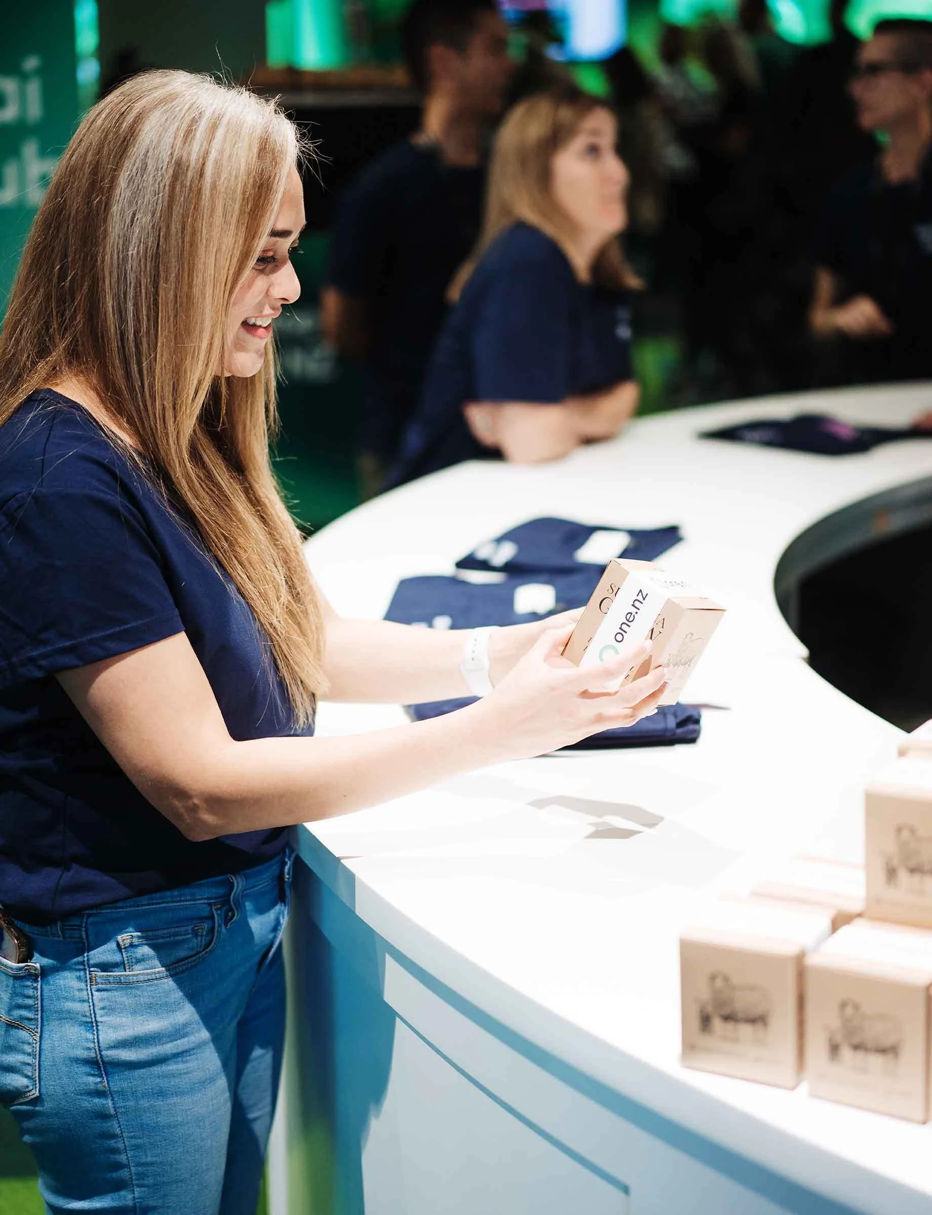 A woman with long blonde hair smiling and looking at a smartphone at a booth with other women in the background at a tech or retail event.
