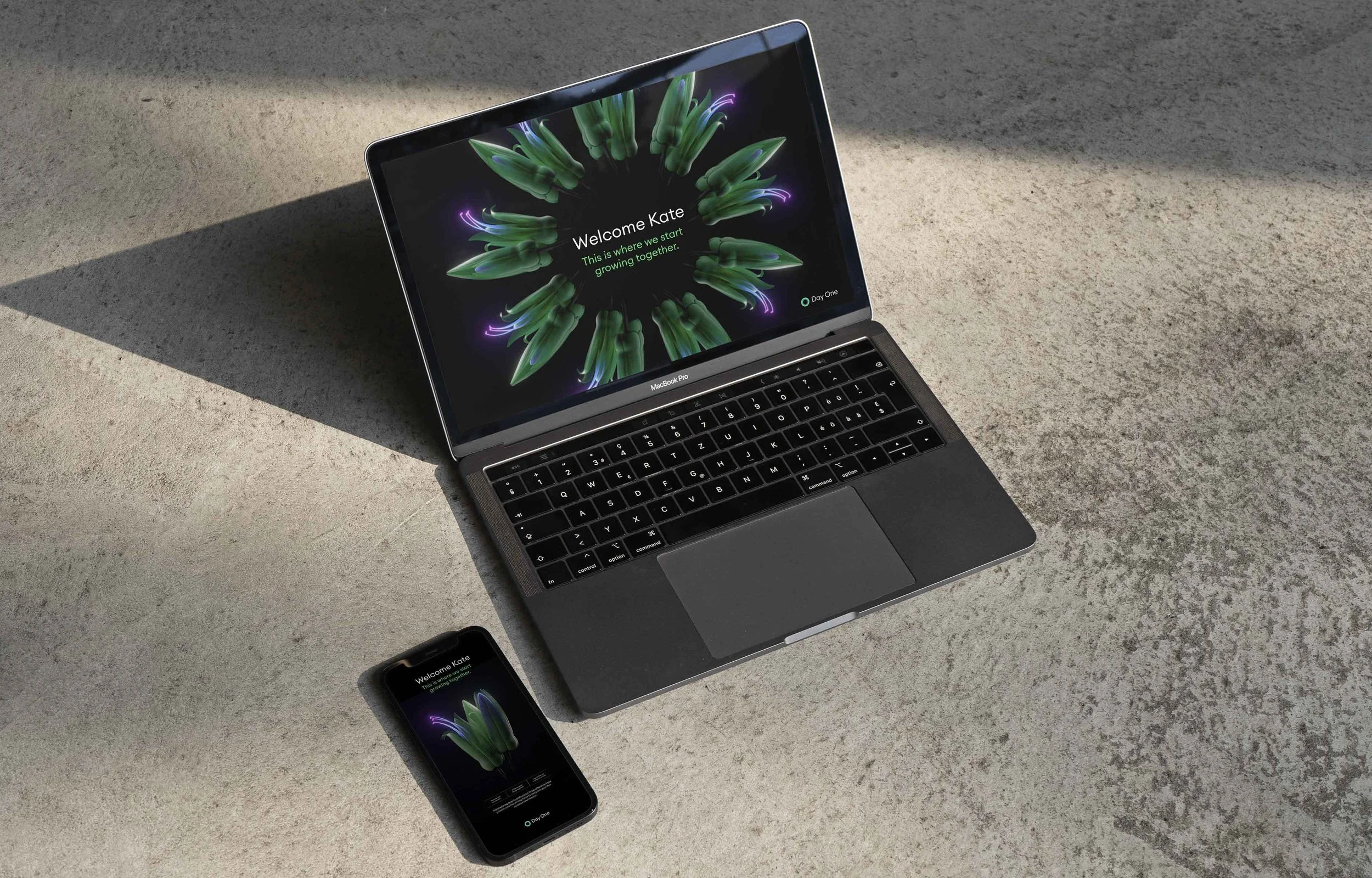 A MacBook Pro laptop and an iPhone on a textured surface, both displaying a welcome screen with a green plant and purple lightning accents, greeting Kate and inviting her to start growing together.