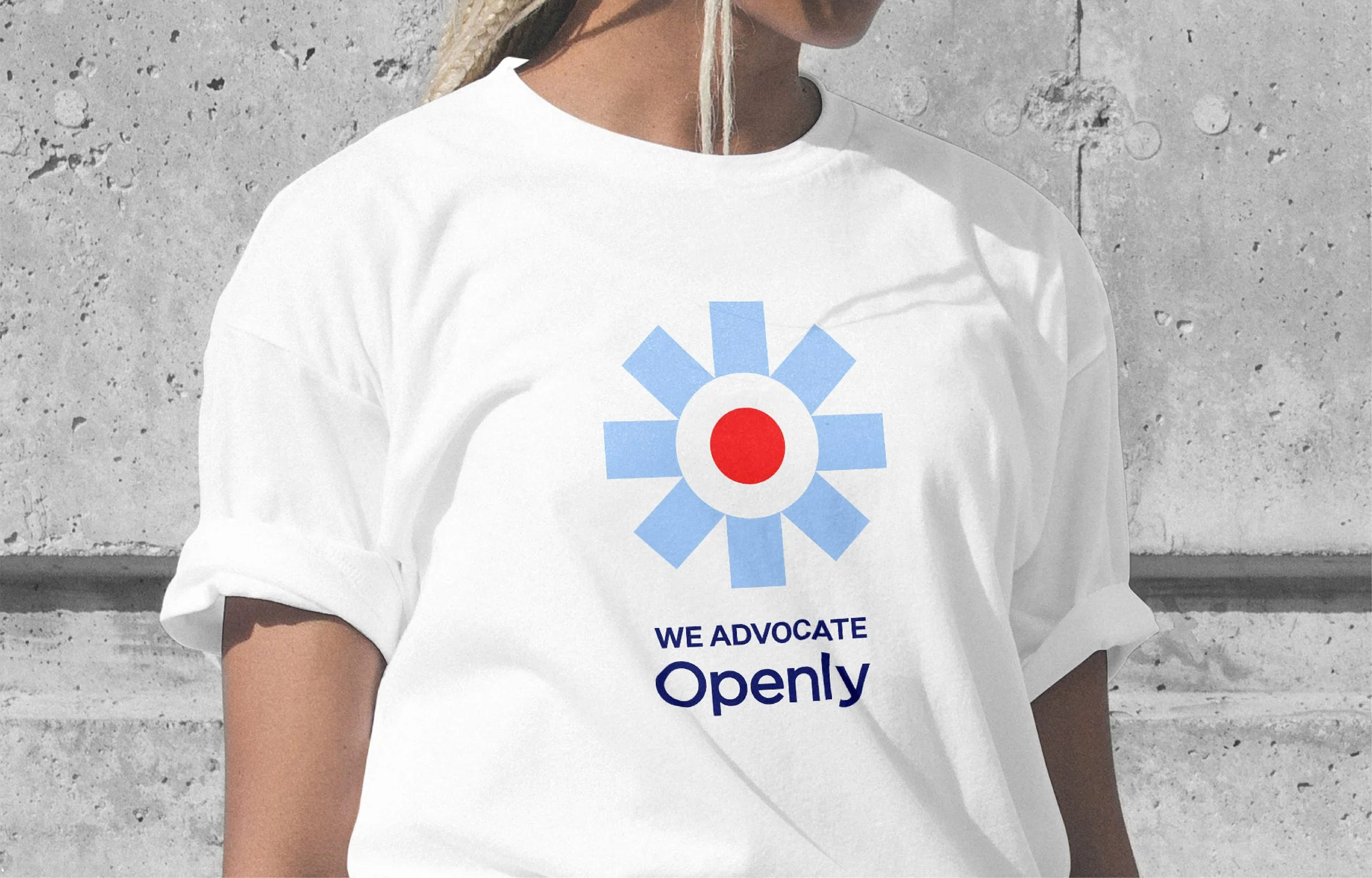 Person wearing a white T-shirt with a graphic of a flower composed of a red circle and blue petal-like shapes, along with the texts "We Advocate" and "Openly" printed below the graphic.