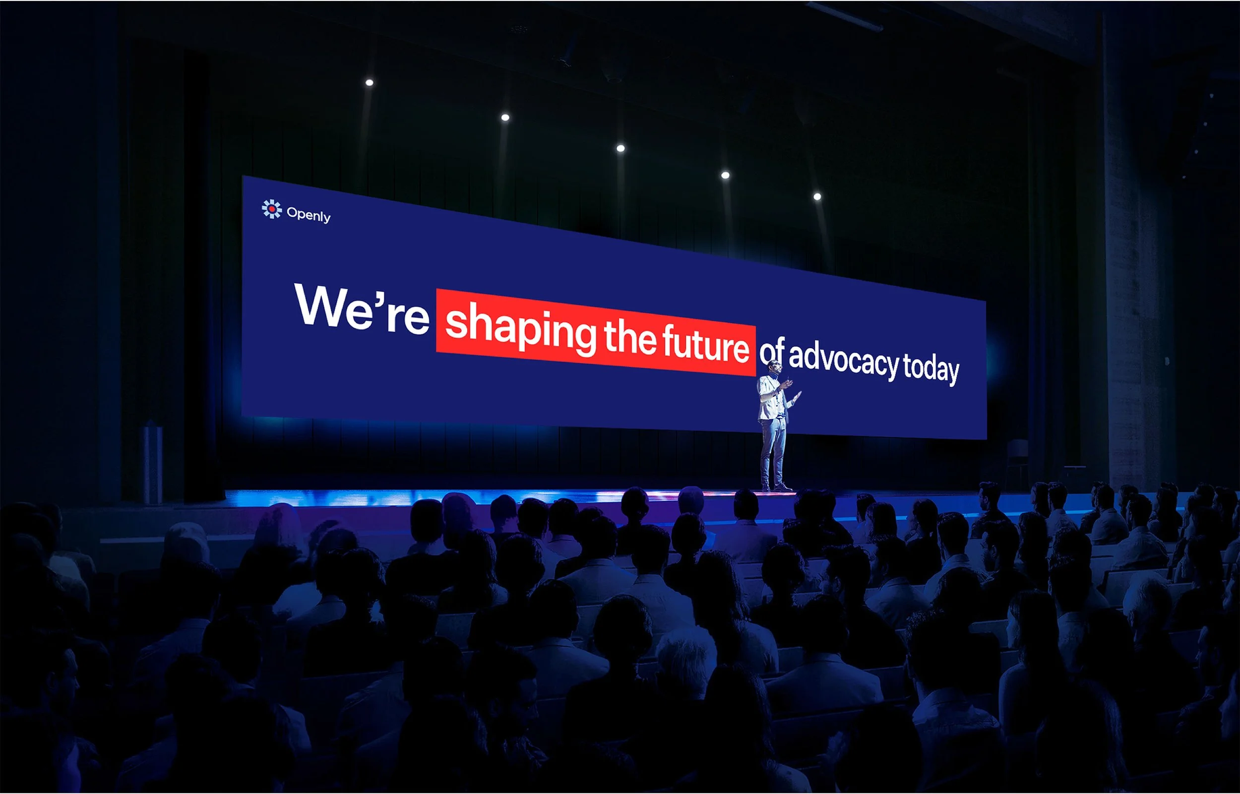 A person speaking on a stage with a large screen behind them displaying the text "We're shaping the future of advocacy today" at a conference or presentation setting.