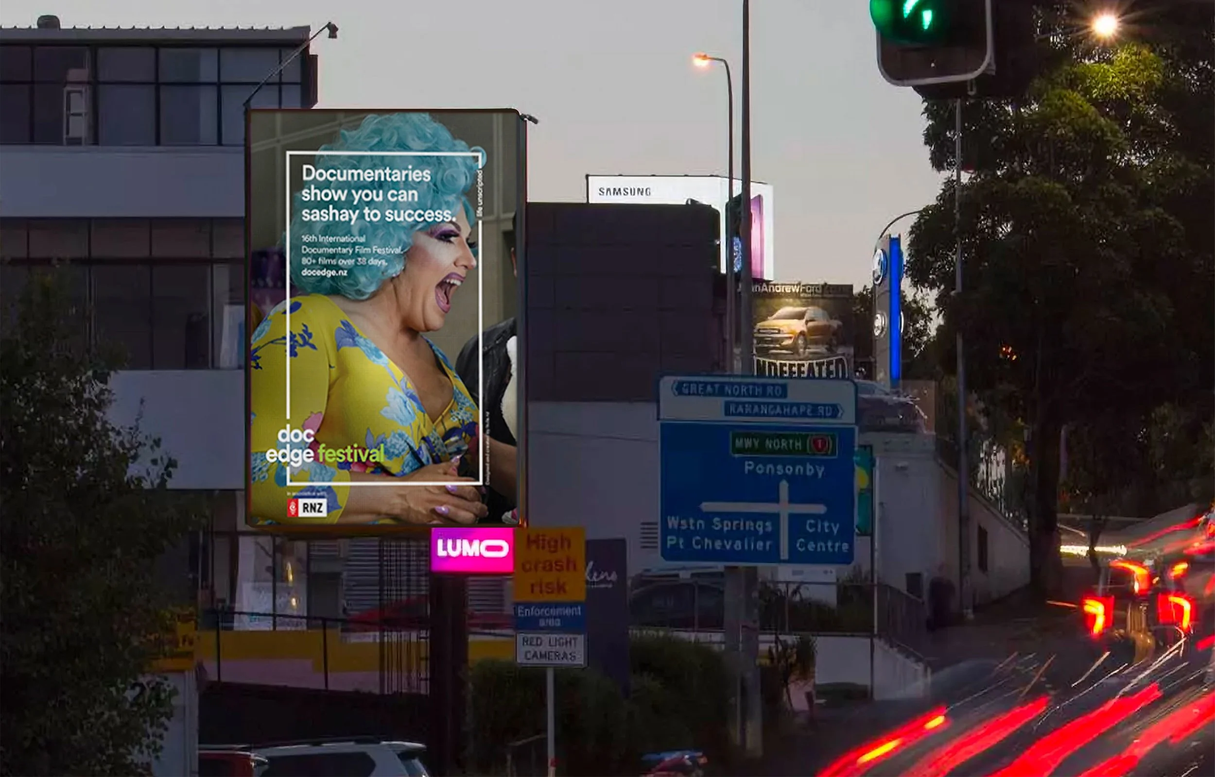 A large billboard advertising documentaries at the Doc Edge Festival, featuring a woman with blue hair and colorful clothing. The scene is at dusk with vehicle lights and street signs visible.