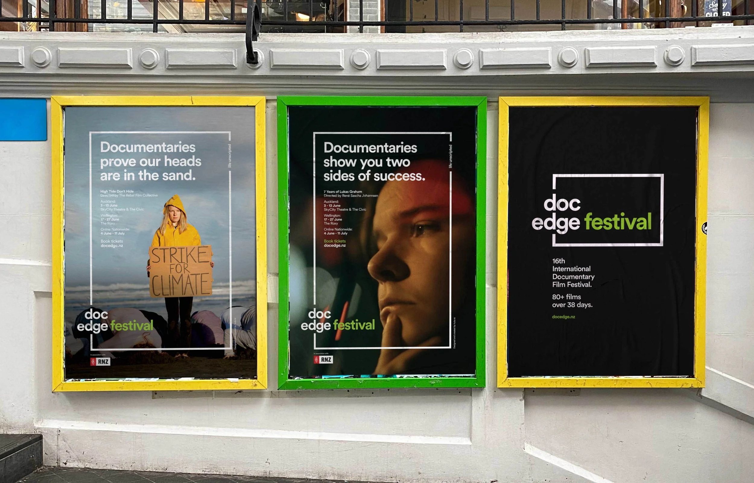 Three posters on a wall promoting the Doc Edge documentary film festival, with each poster framed in yellow, green, and yellow respectively.