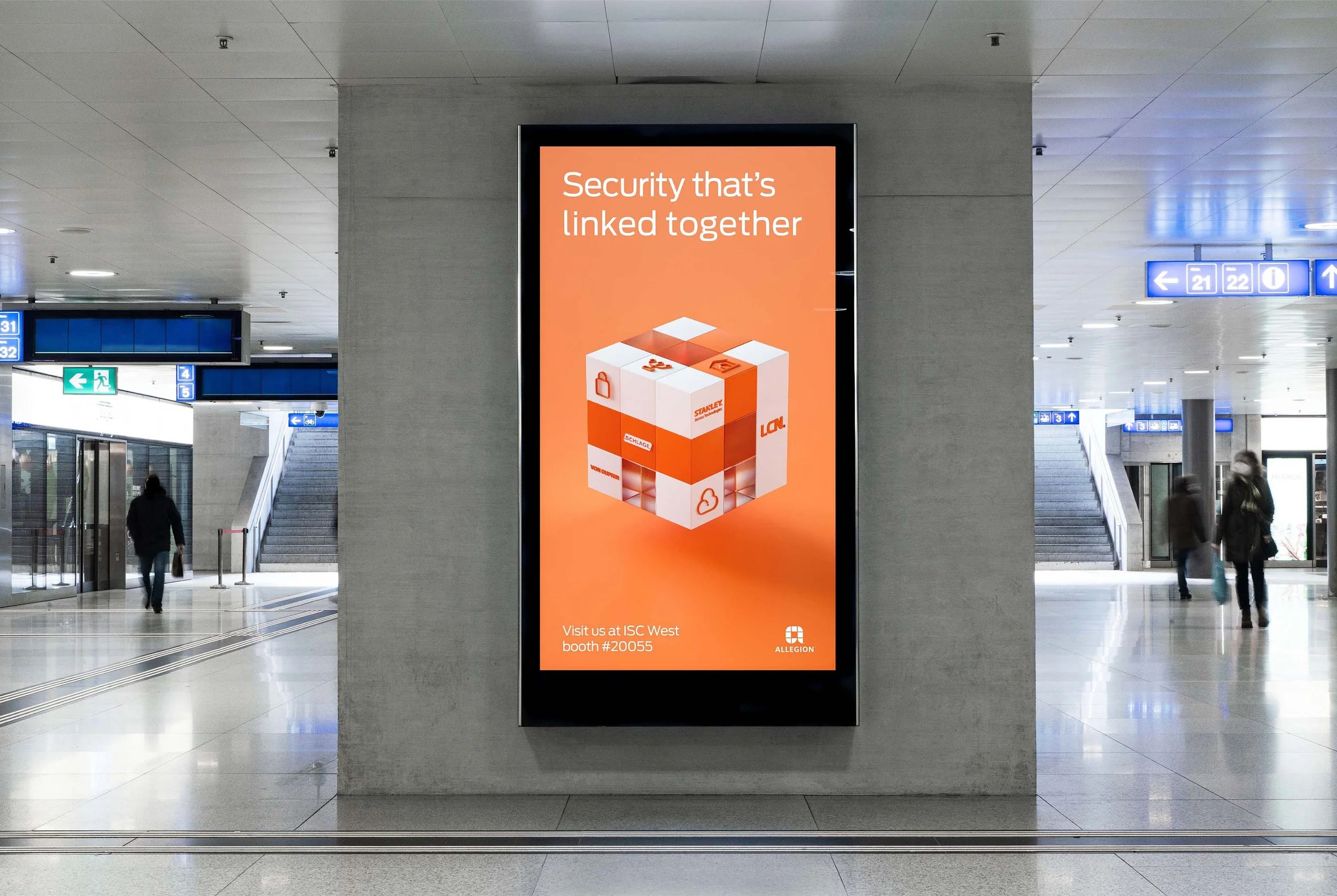 Digital advertisement on a lit display screen in a train station, promoting security linked together, featuring a 3D cube with security icons and text, along with visit details and company branding.