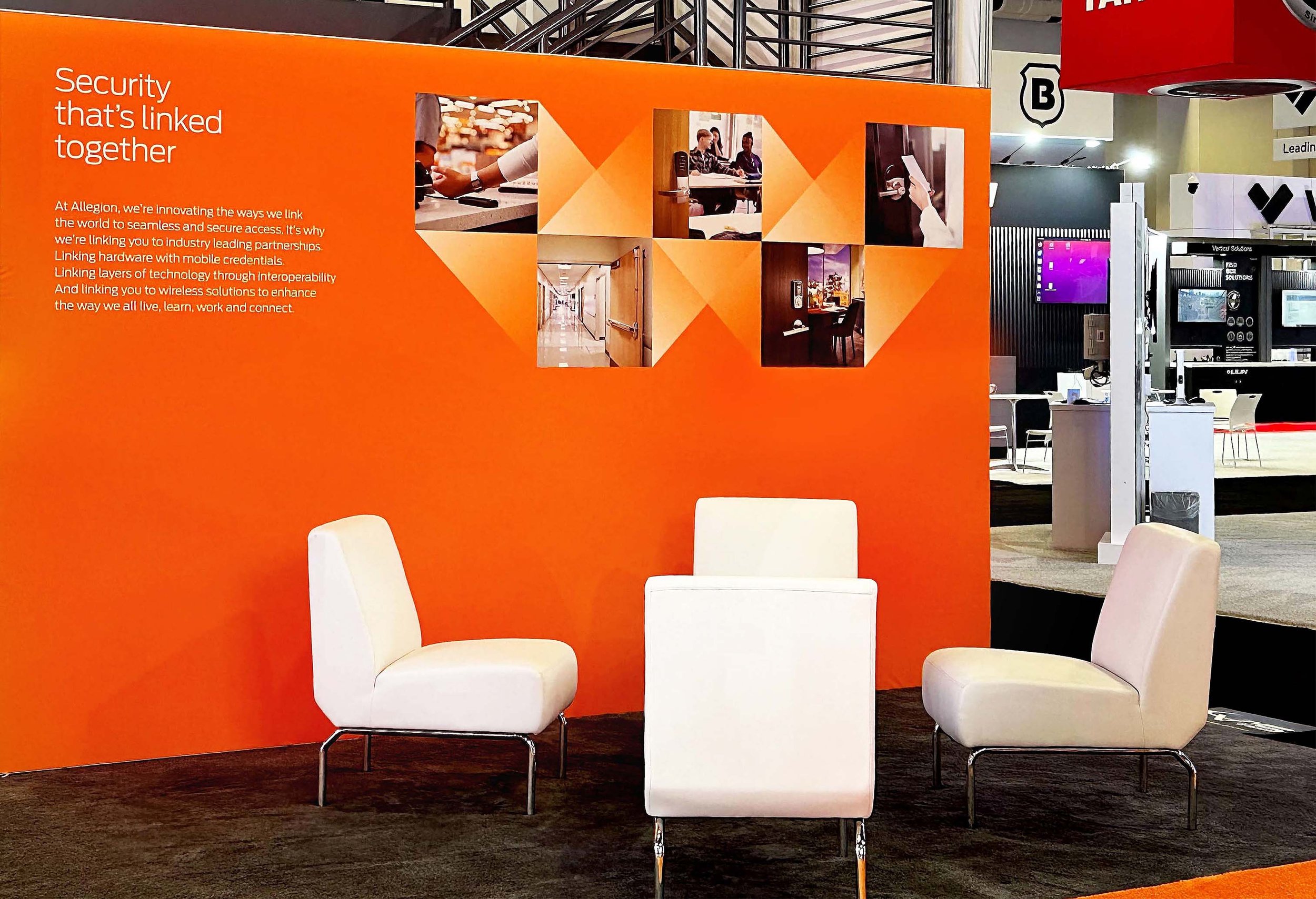An orange exhibition booth with white chairs and a white panel table in front. The booth has text that reads 'Security that's linked together' and a paragraph about linking security technologies and solutions. There are four photographs on the upper part of the booth showing people working on laptops, in meetings, and walking in a hallway.