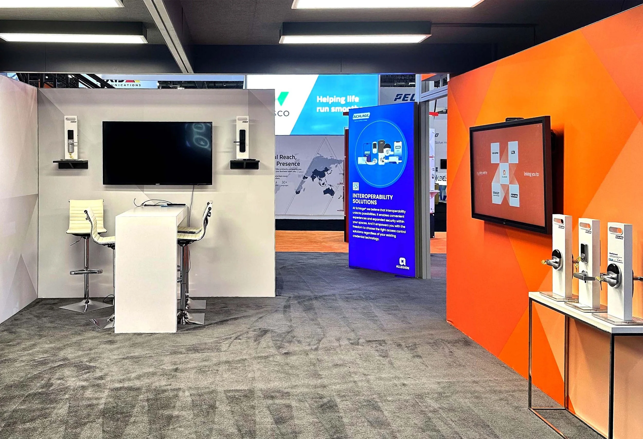 Trade show booth with a large display screen, a smaller screen, a table, and electronic devices on stands. The booth features white and orange walls, with informational signage about interoperability solutions.